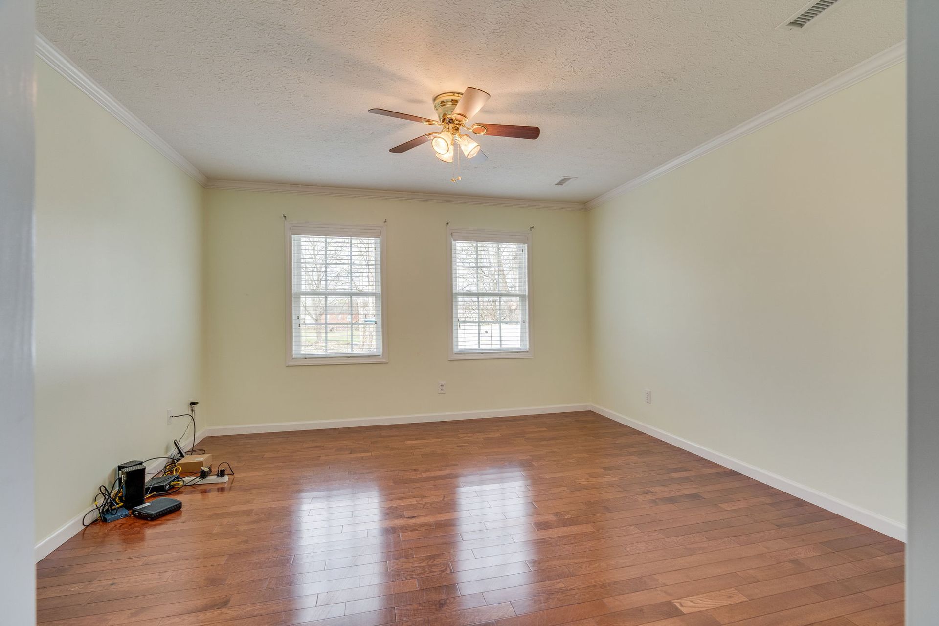 An empty room with light yellow walls, wood flooring, a ceiling fan, and two windows facing outward.