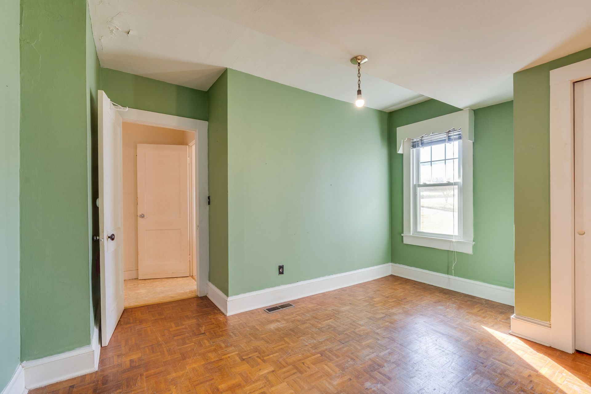 Empty room with green walls, white trim, and hardwood floors. A doorway leads to another room. A window is present.