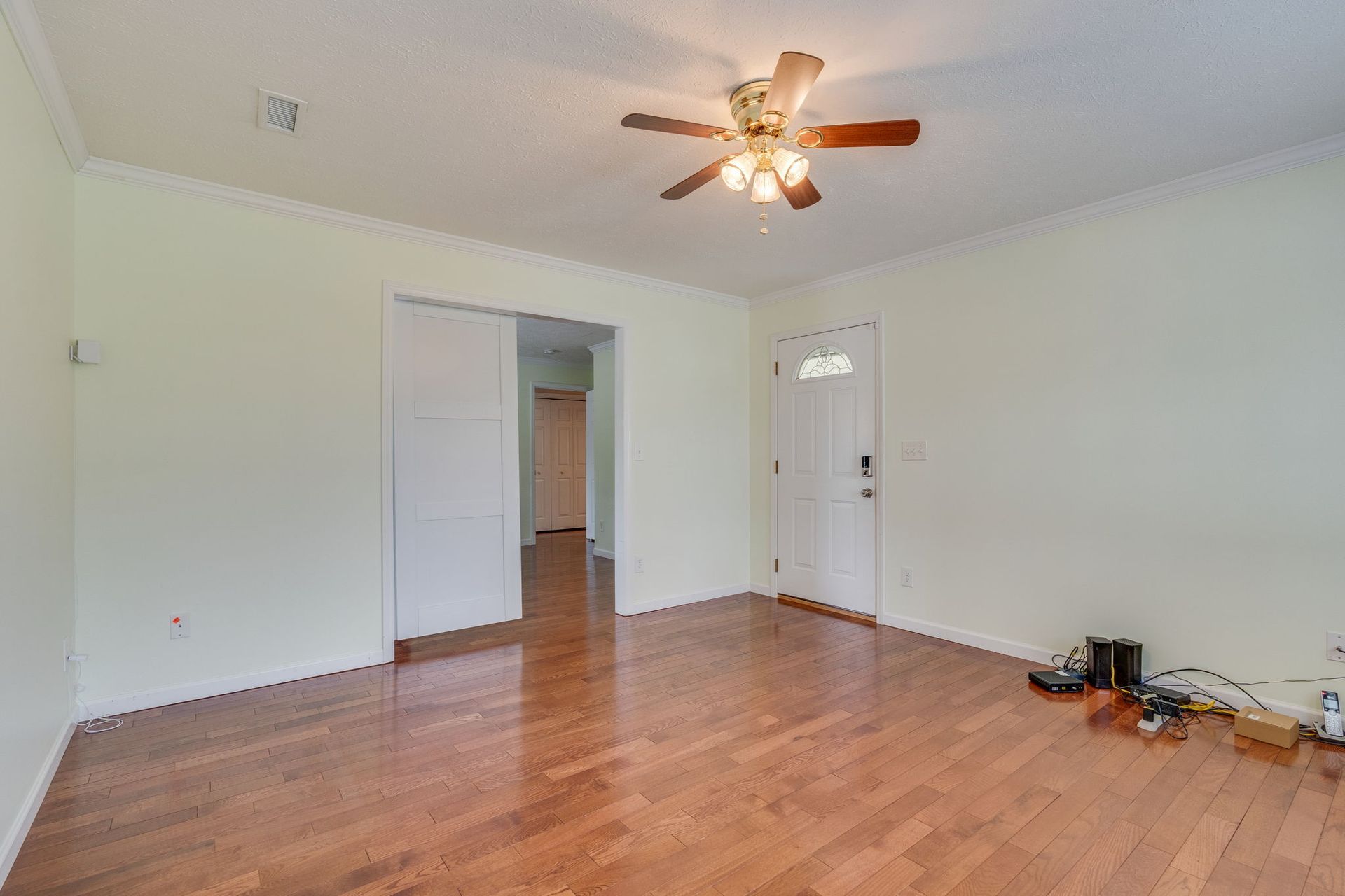 An empty room with light walls and polished hardwood floors, featuring a ceiling fan and an open doorway.