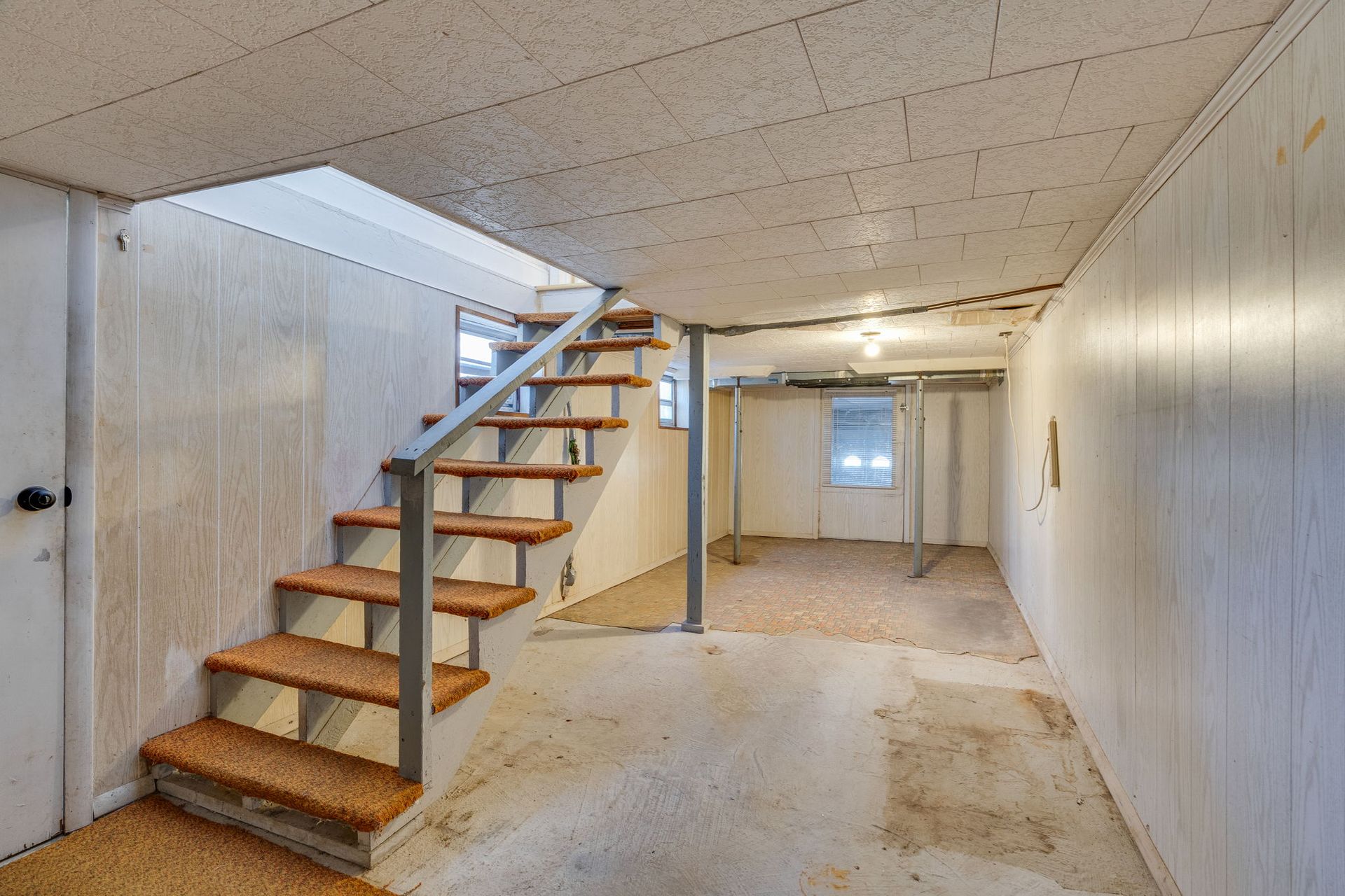 Basement interior with stairs leading up, white panel walls, and concrete floor.