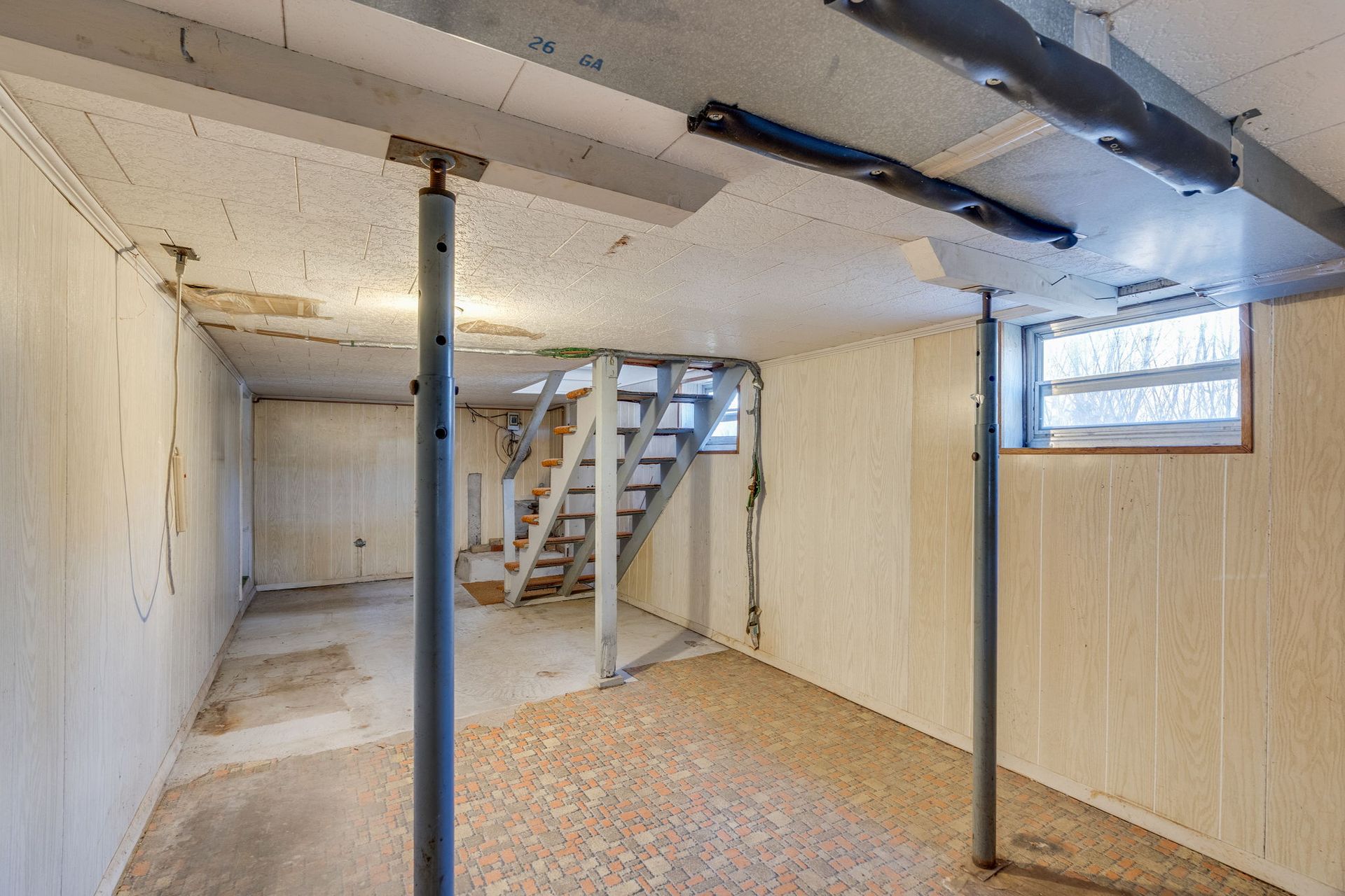 Basement with support beams, stairs, and a small window. White paneling and concrete floor.