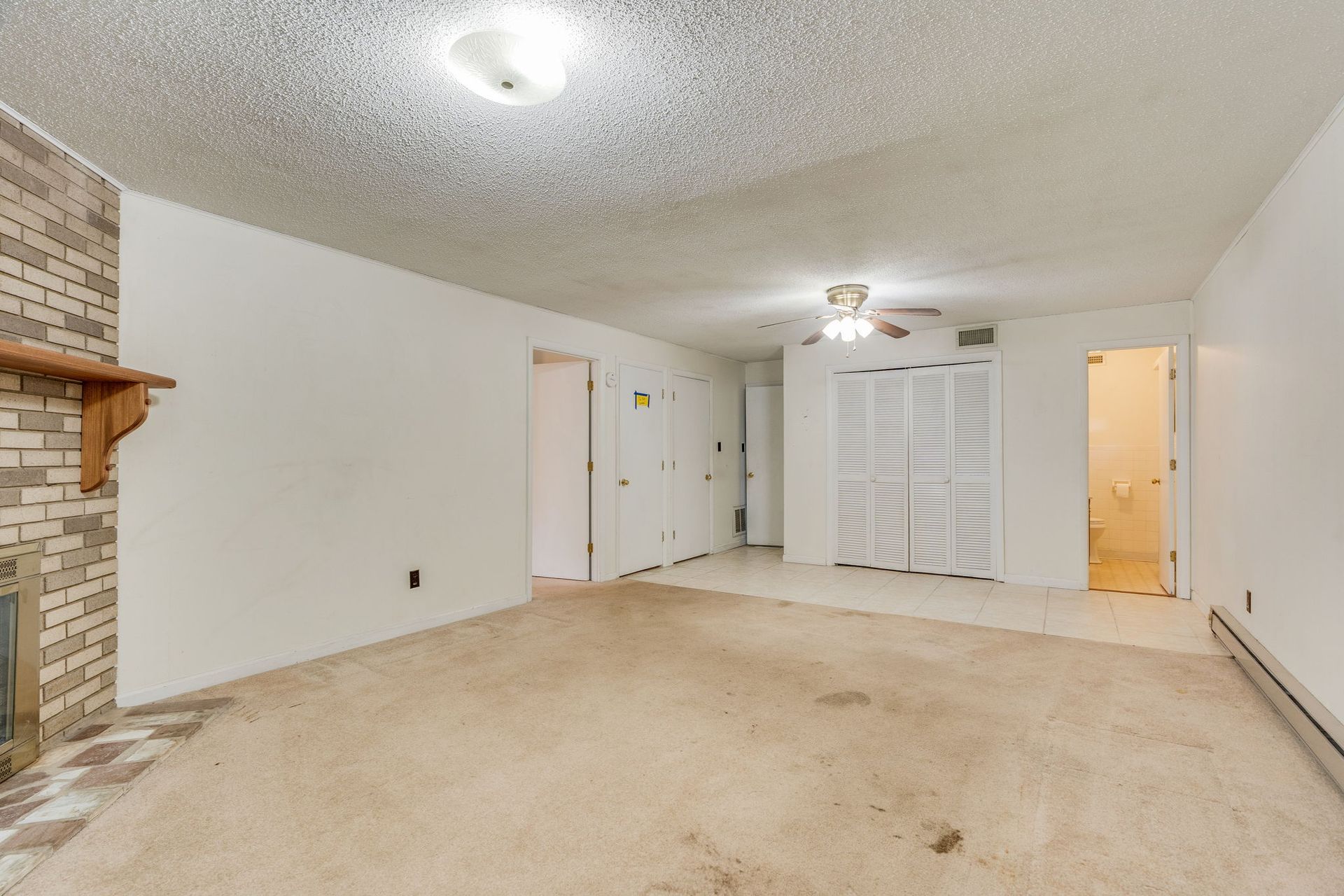 An empty room with tan carpet, white walls, a brick fireplace, ceiling fan, and doors leading to a hallway and bathroom.