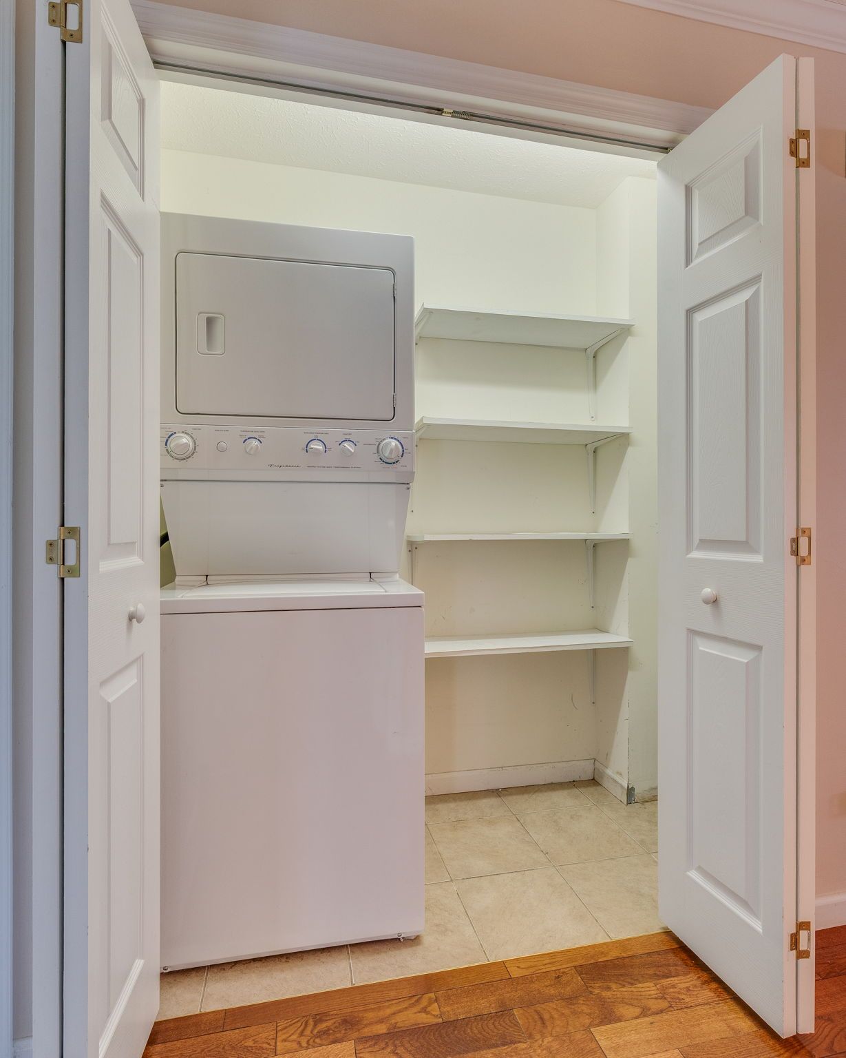 A stacked white washer and dryer unit inside a closet next to four empty white shelves.