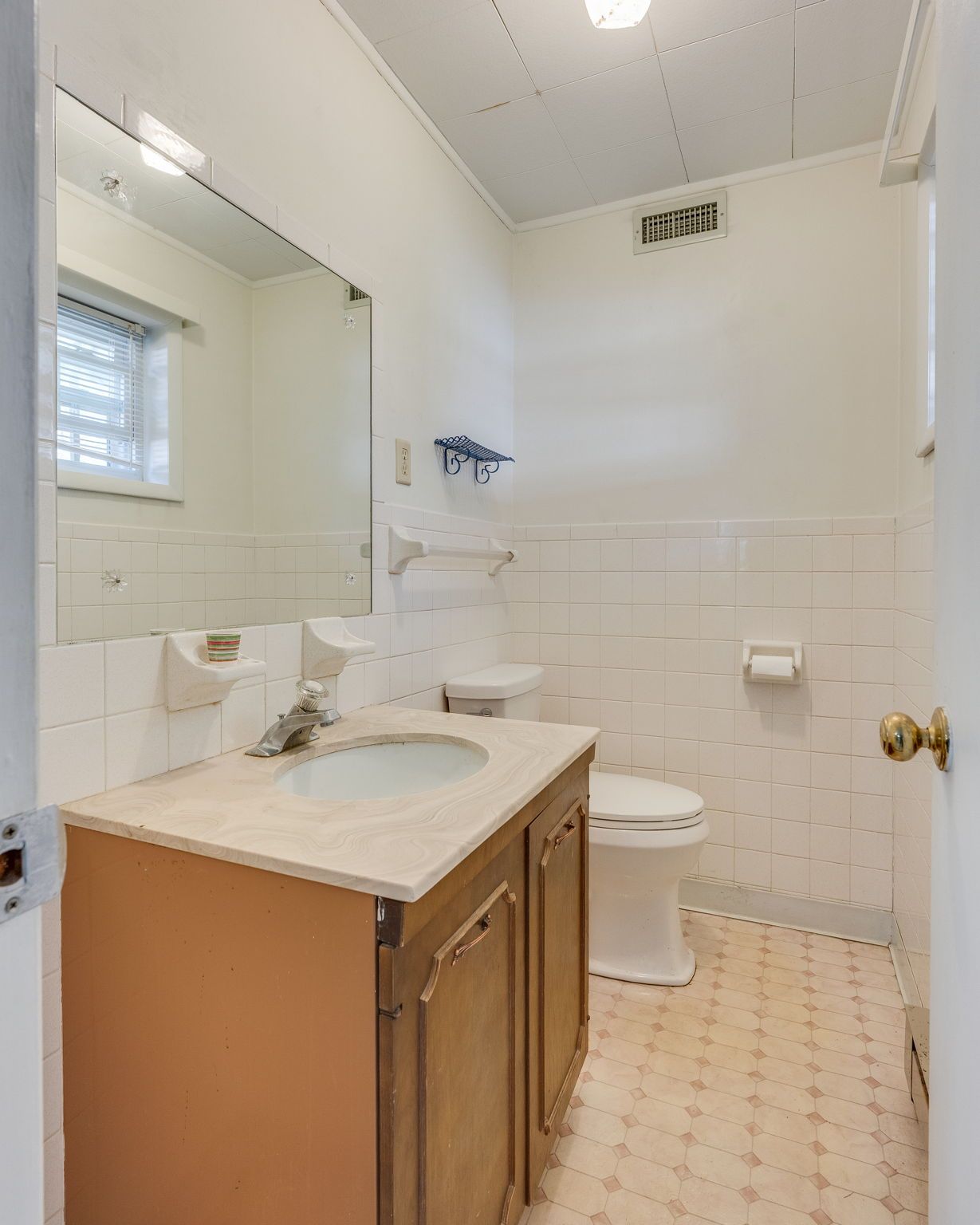 A bathroom with white tiled walls, a wooden vanity with a white countertop, a mirror, and a toilet.