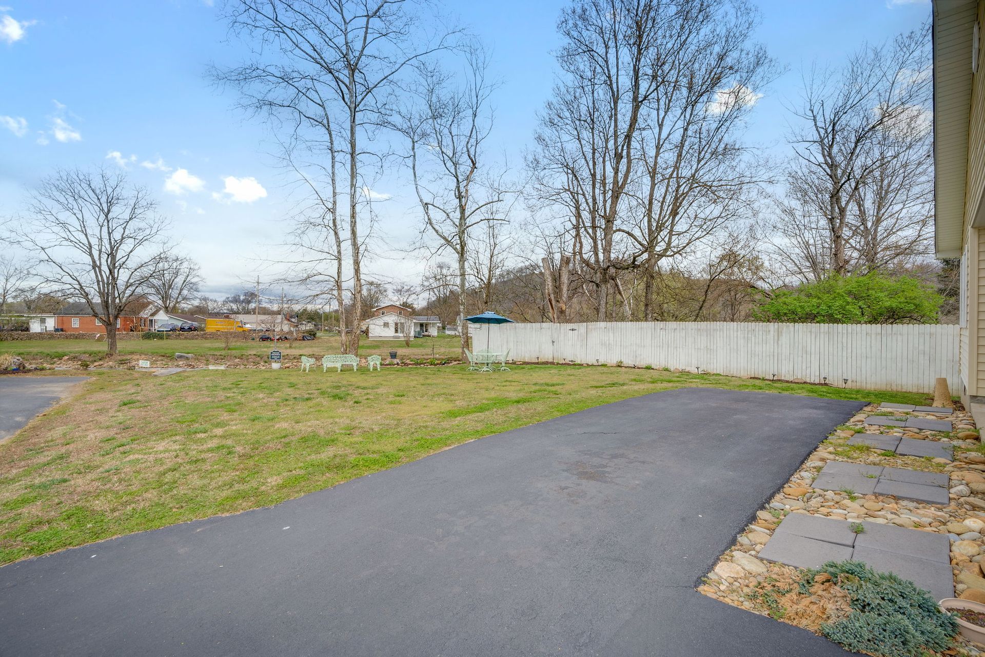 An asphalt driveway leads toward a grassy lawn and white fence with trees under a blue sky.