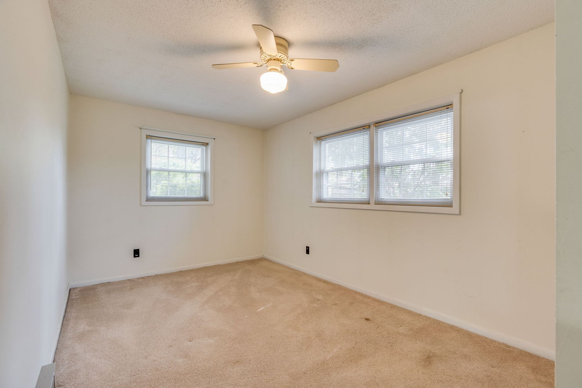An empty bedroom with beige carpet, cream walls, a ceiling fan, and two windows with white blinds.