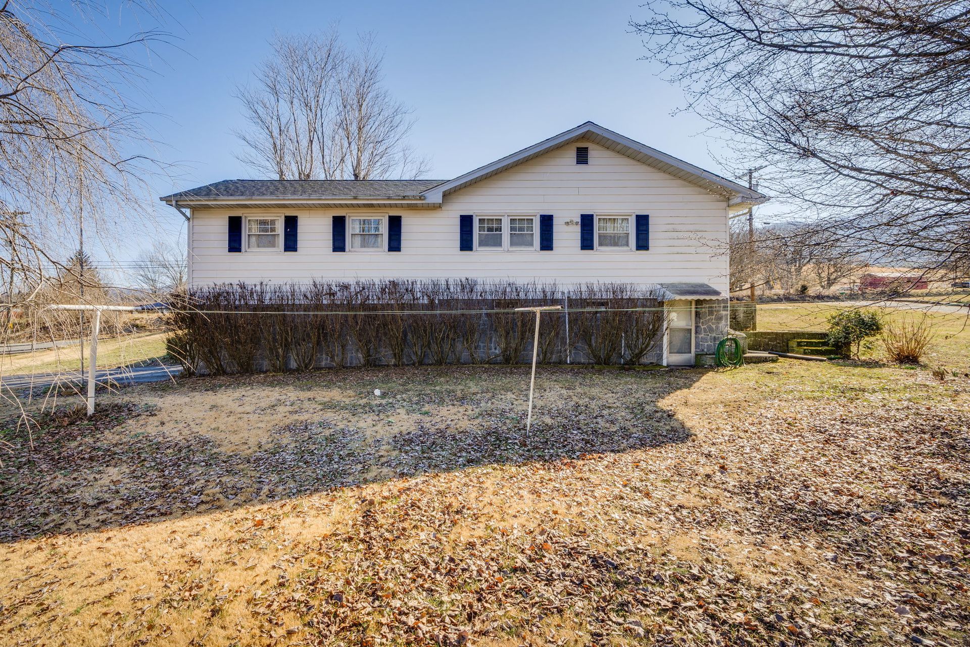 Two-story white house with blue shutters, surrounded by bare trees and dry leaves in a sunny outdoor setting.