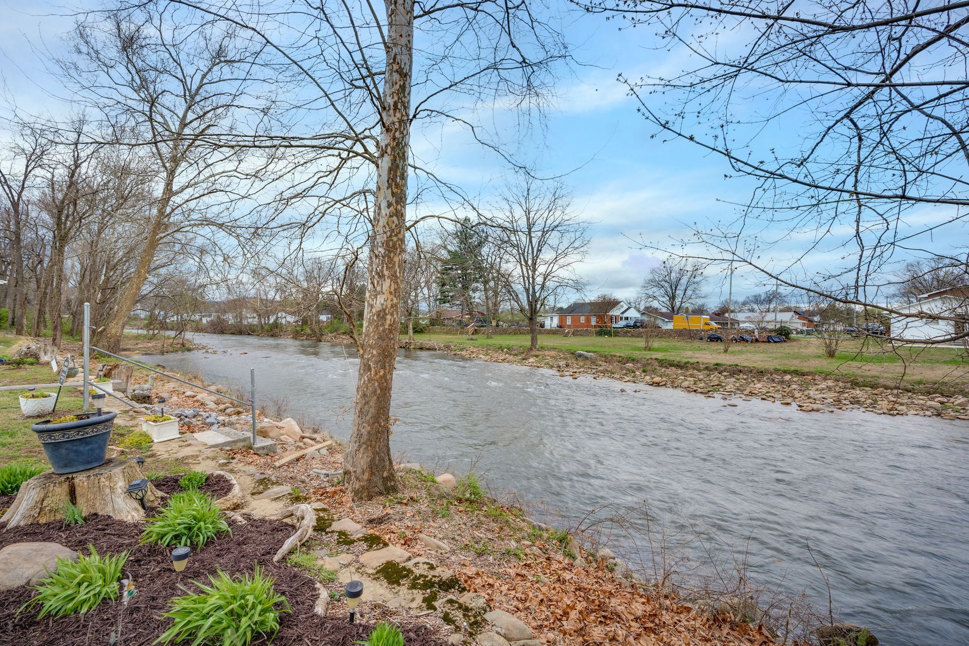 A stream flows through a grassy landscape lined with bare trees, rocks, and small plants under a cloudy blue sky.