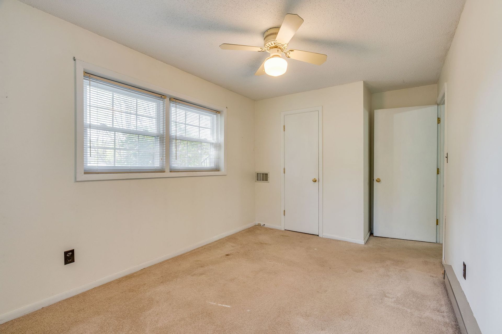 An empty, neutrally colored bedroom with tan carpet, a ceiling fan, a window, and two white doors.