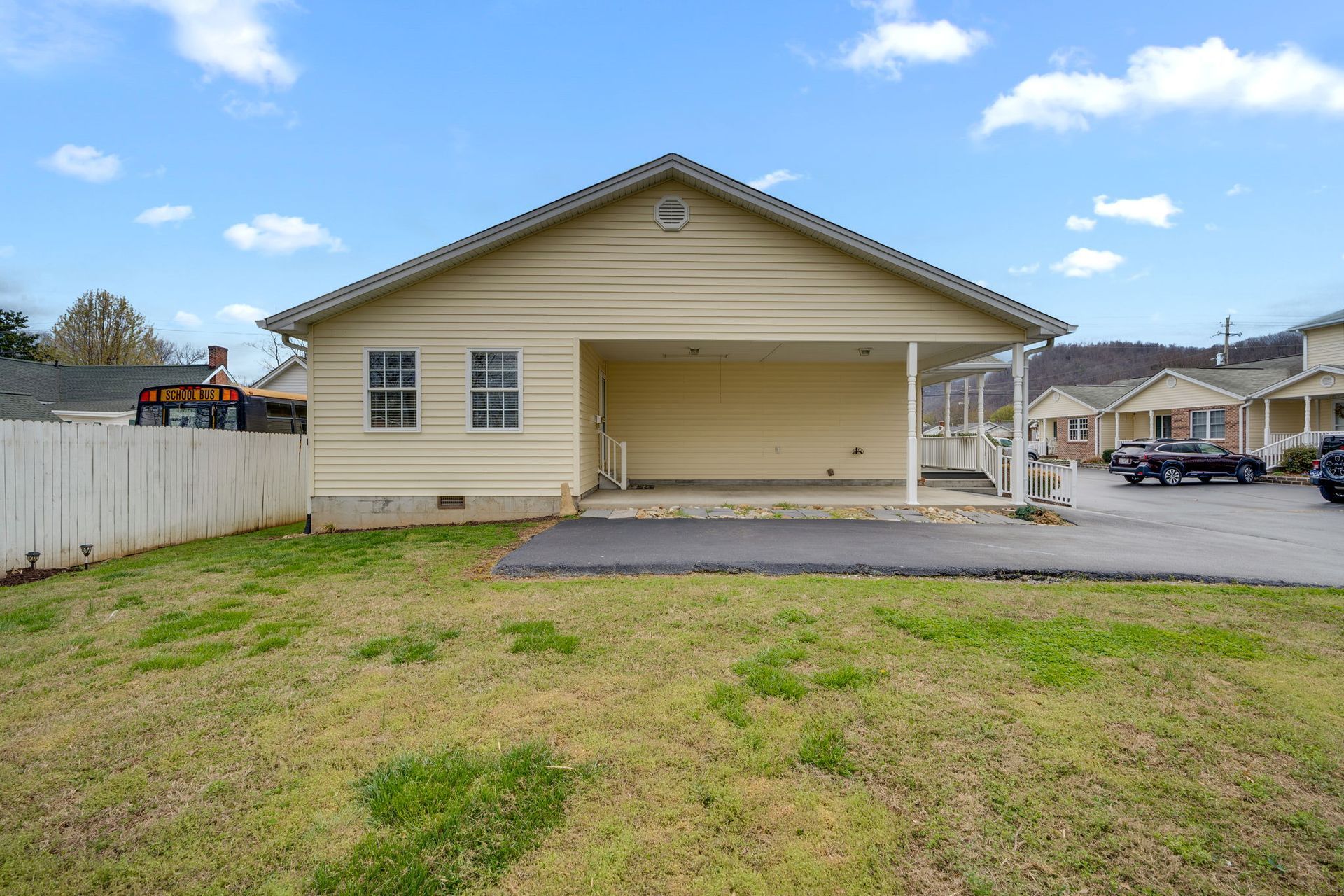 A light yellow house with a covered carport, seen from a grassy lawn under a blue, partly cloudy sky.