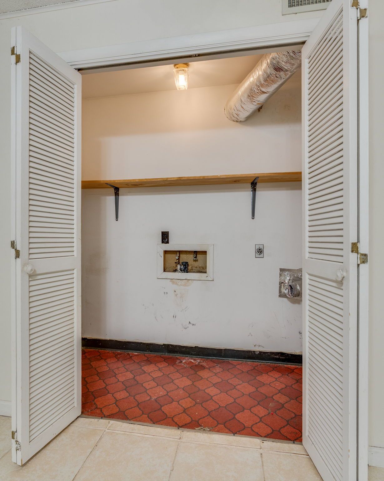A laundry closet with red tile floors, a wooden shelf, a light fixture, and plumbing hookups behind white louvered doors.