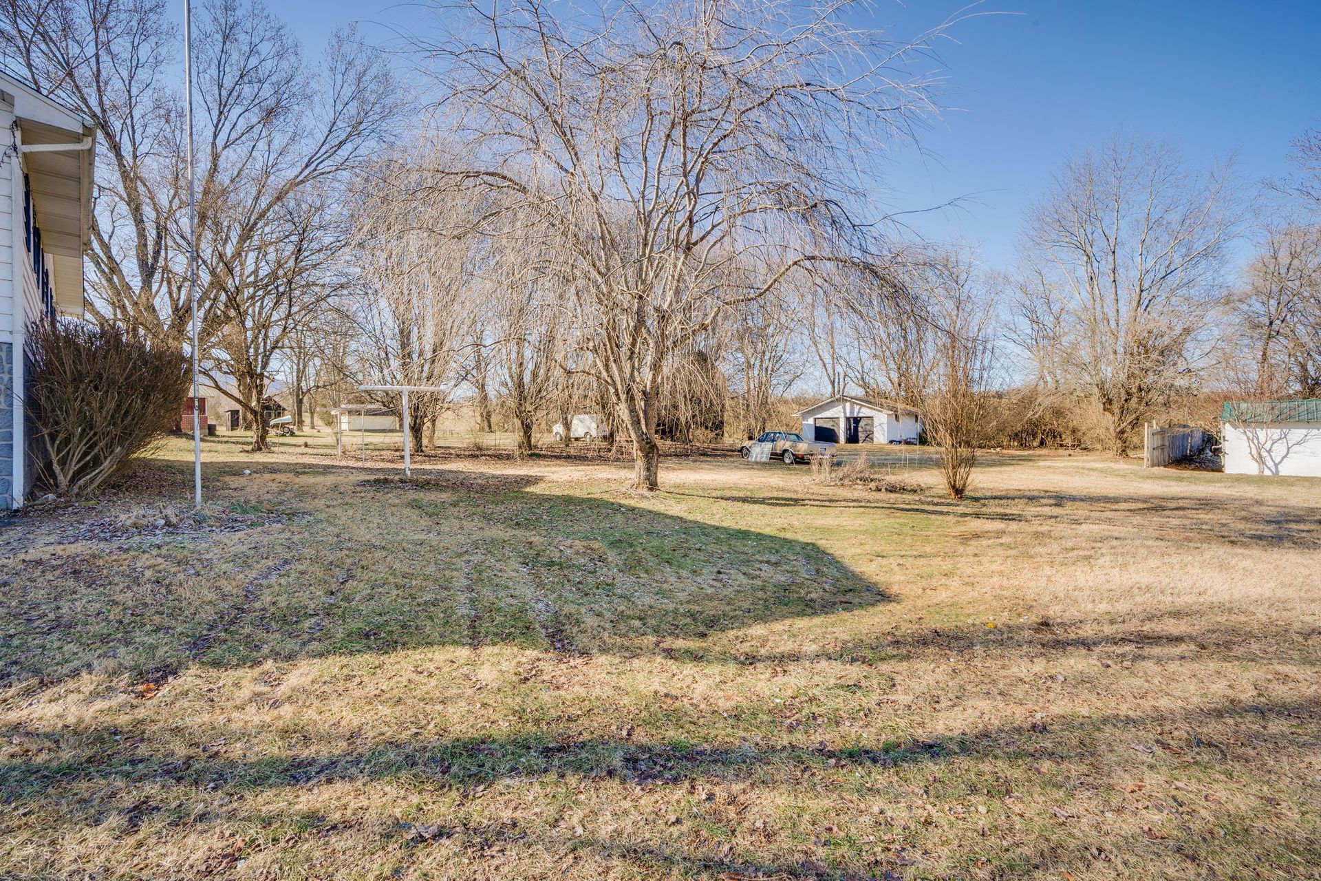 Grassy backyard with bare trees, shed, and small buildings under a blue sky.