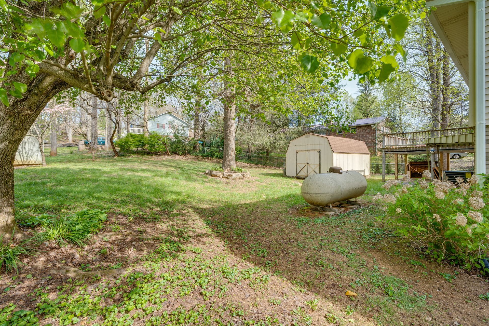 A grassy backyard featuring a white shed, a large propane tank, mature trees, and a wooden deck attached to a house.