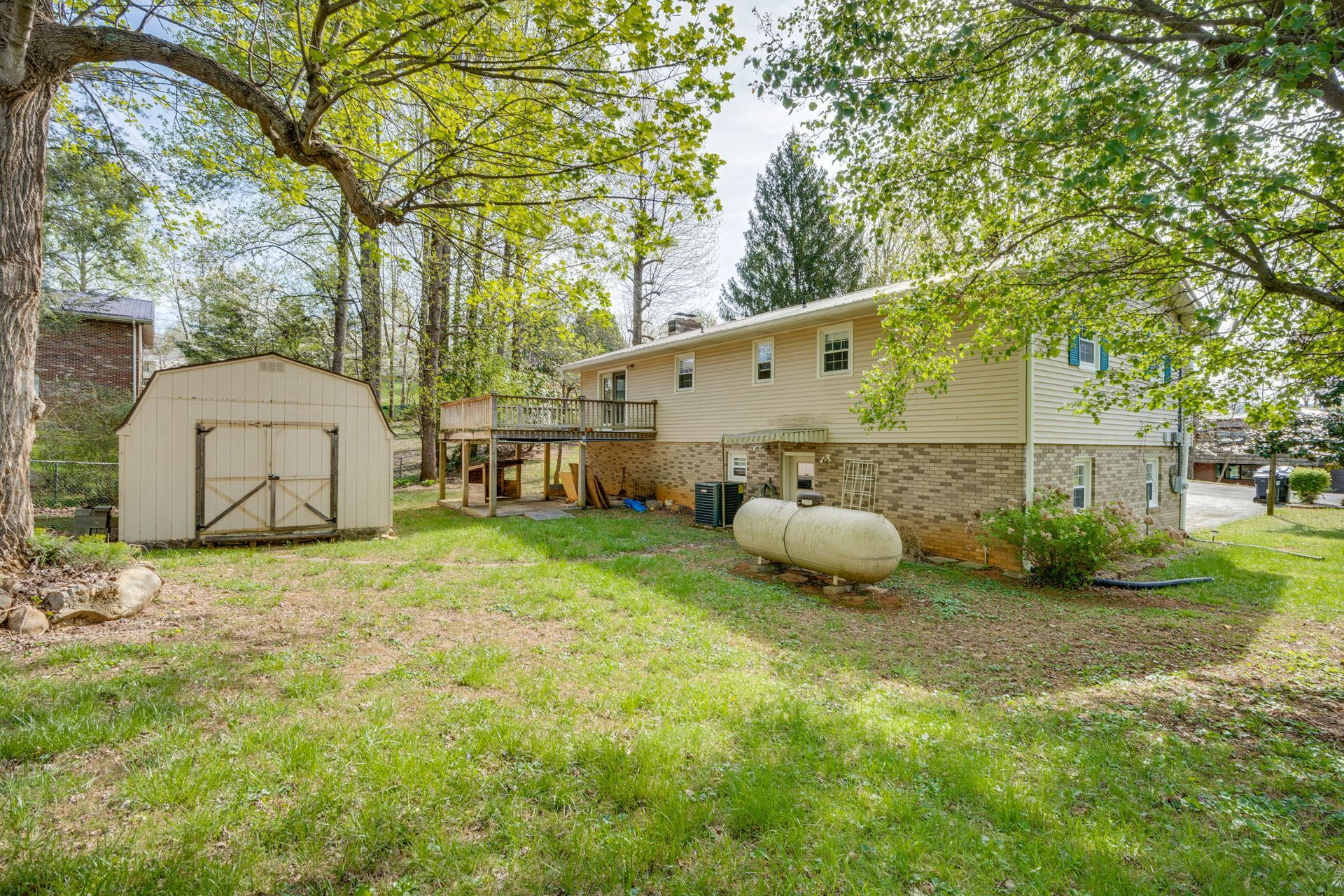 A beige house with a brick base and a shed in a grassy backyard, surrounded by trees on a sunny day.