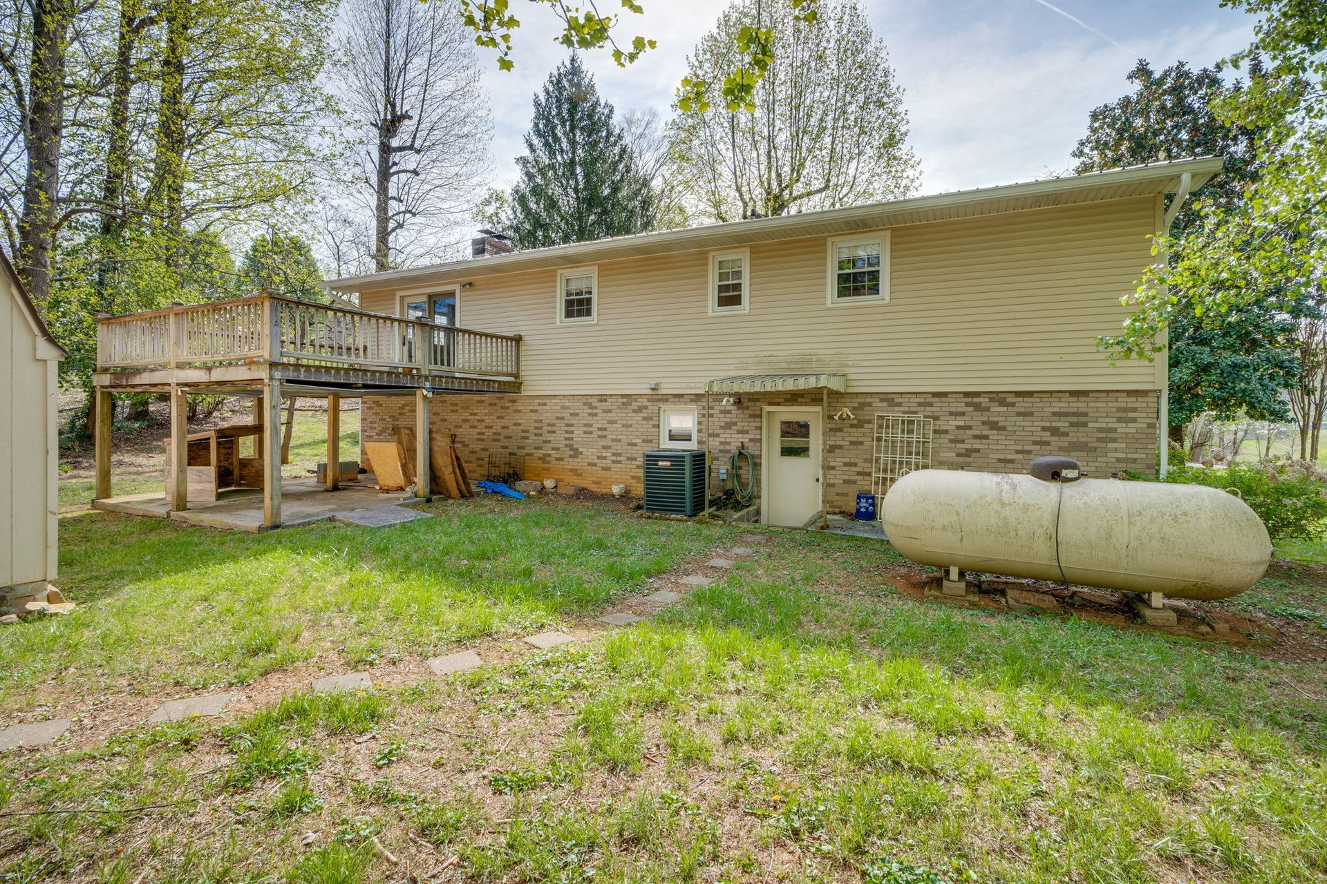 Back exterior of a two-story house with tan siding, stone foundation, wooden deck, and a large propane tank on the lawn.