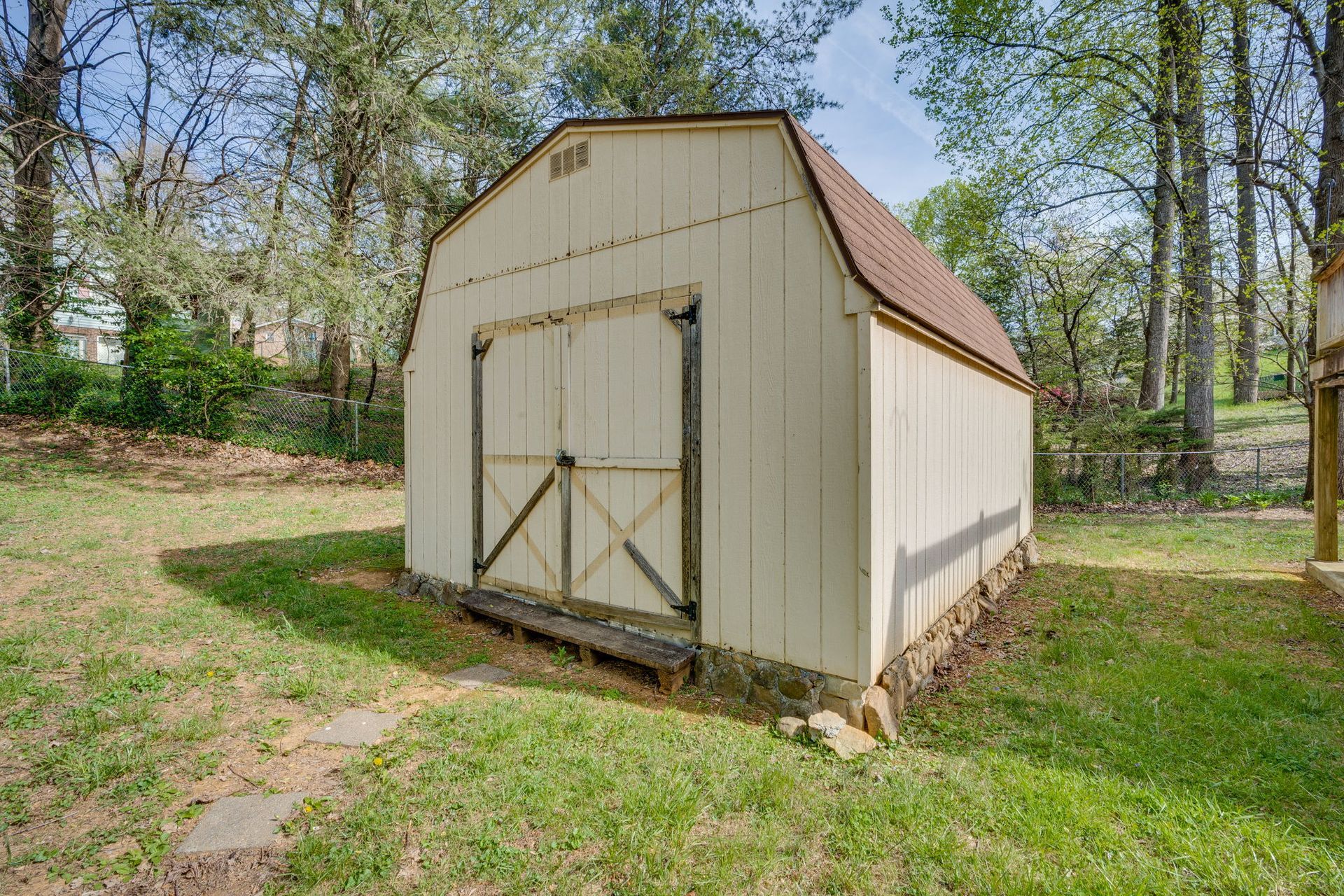 A light beige wooden storage shed with a gambrel roof sits on a grassy yard surrounded by trees on a sunny day.