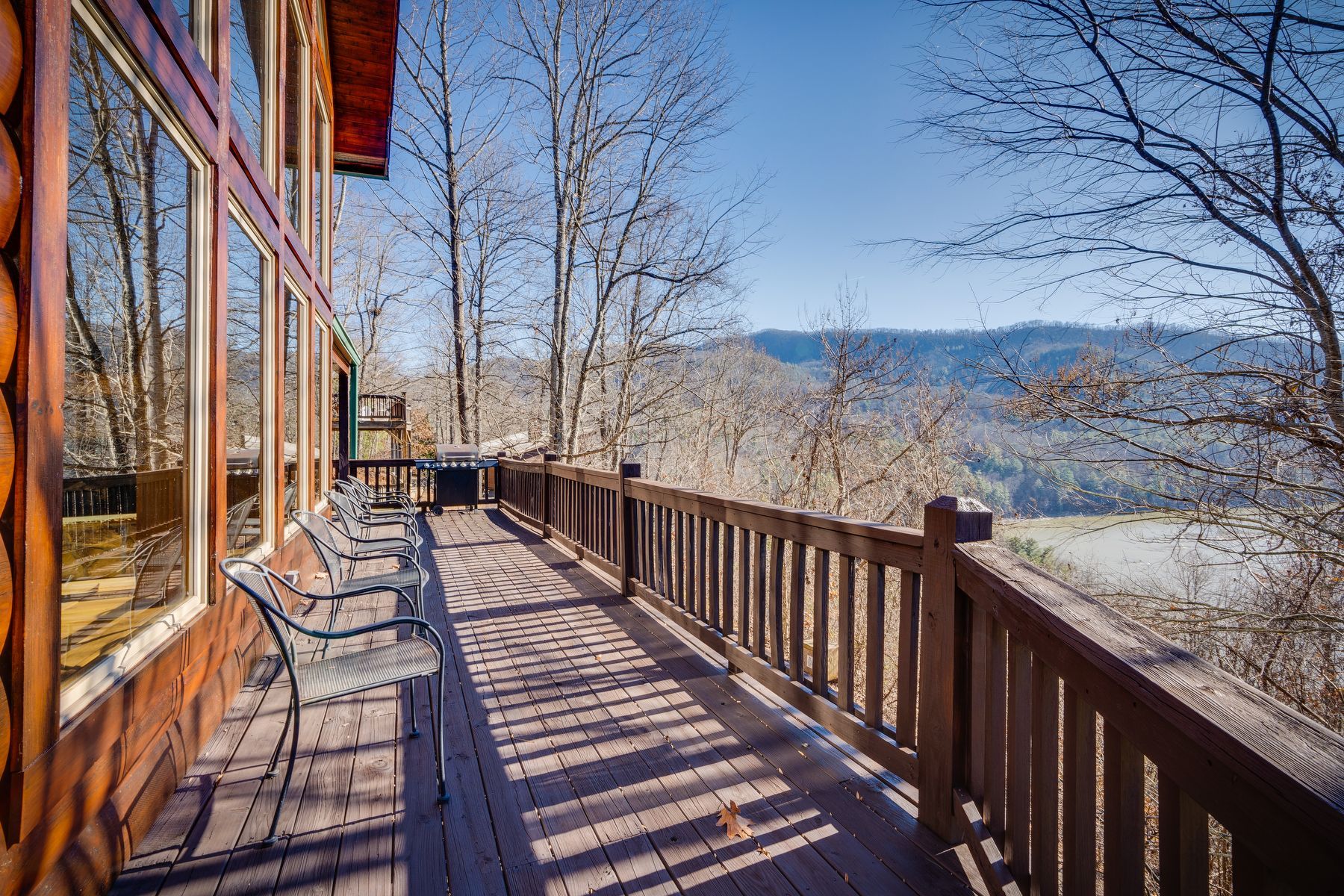 Wooden deck with railing and view of mountains.  Metal chairs sit on the deck near large windows.