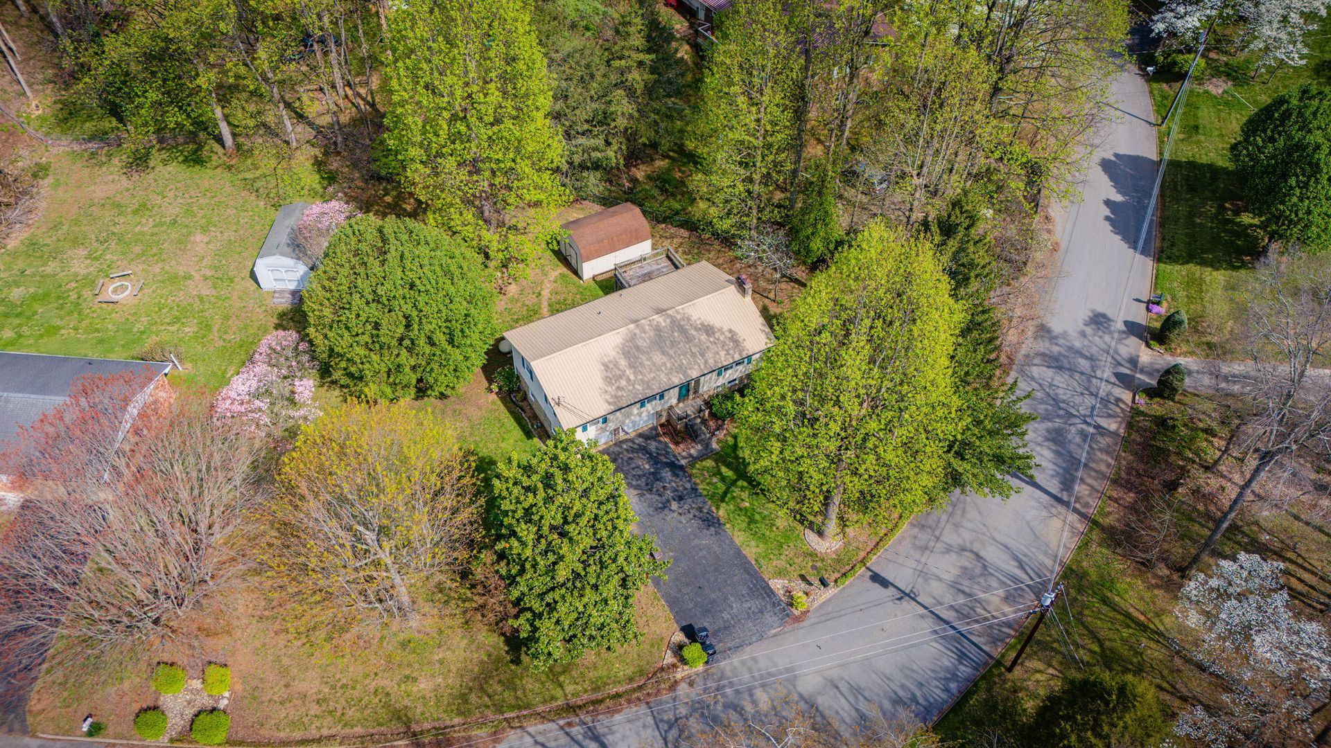 Aerial view of a rectangular house with a light-colored roof, surrounded by trees and a paved driveway in a yard.