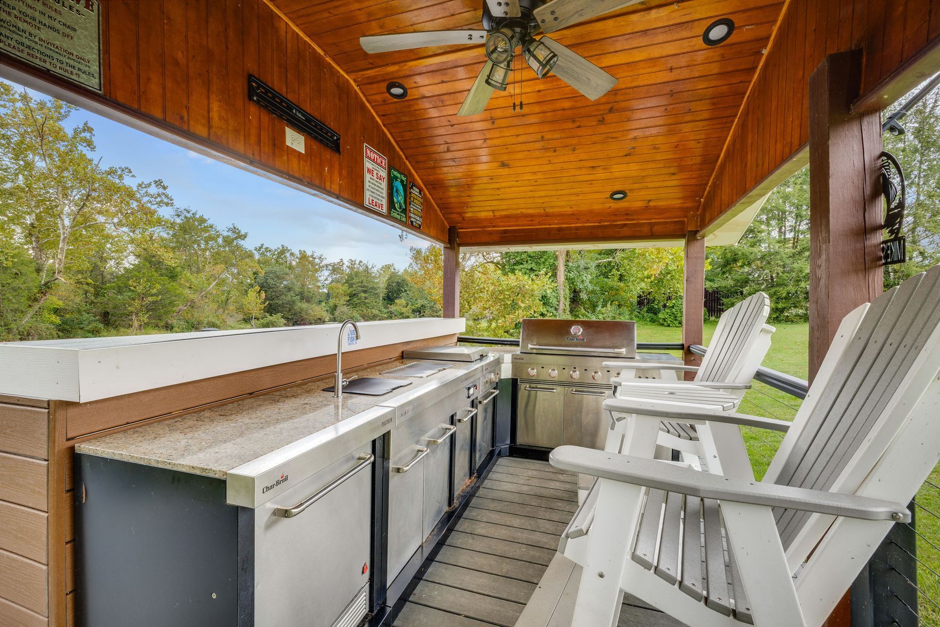 Outdoor kitchen under a wooden roof with grill, sink, and refrigerator, with white chairs and greenery in the background.