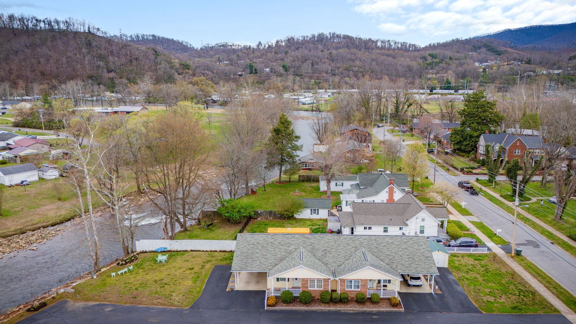 An aerial view of a suburban duplex with a grey roof, dark driveway, and green lawn, located near a creek and hillside.