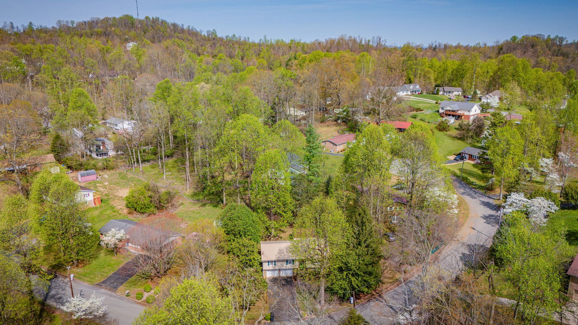 An aerial view of a wooded residential neighborhood featuring several houses nestled among dense green trees and hills.