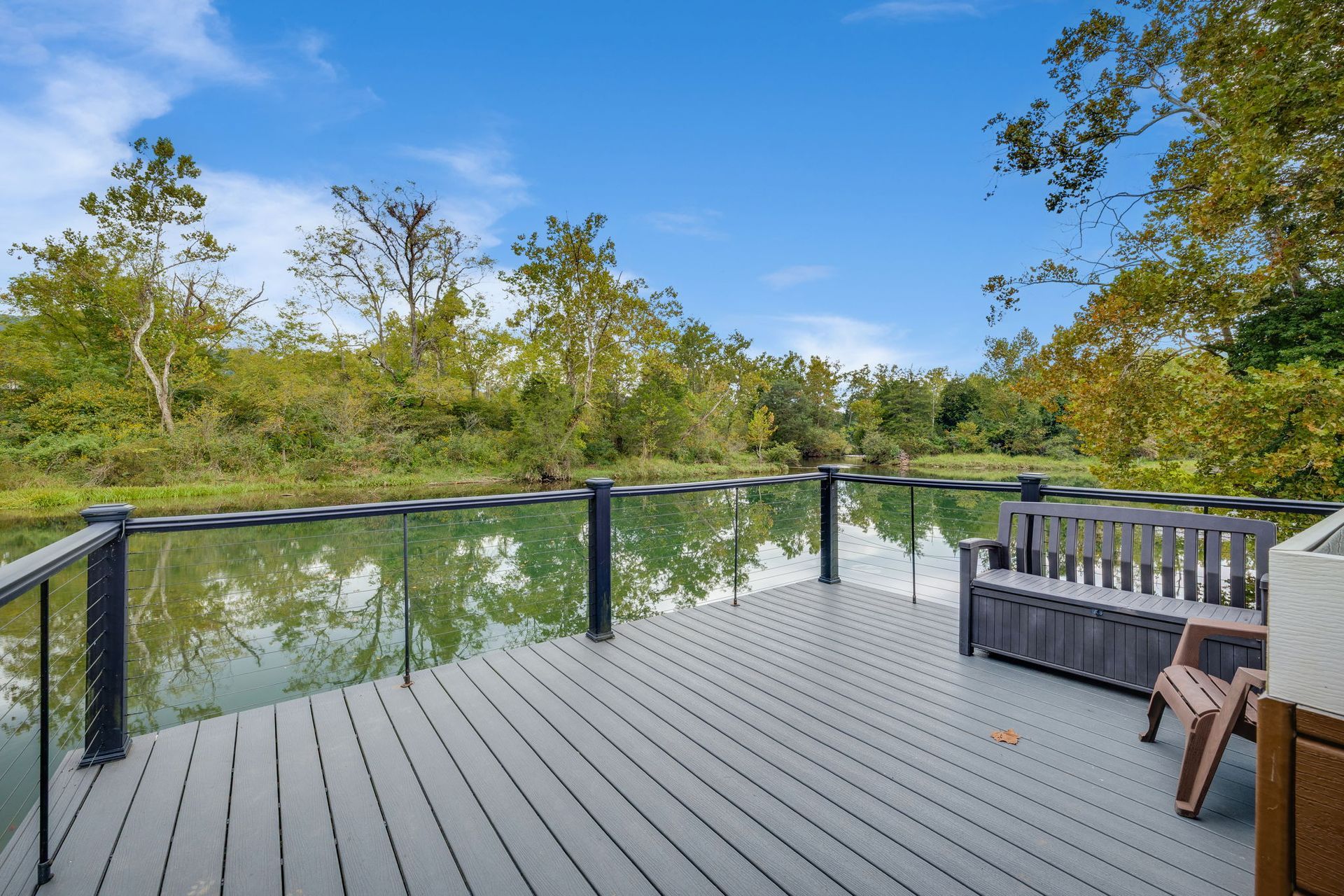 Deck overlooking a calm body of water, featuring a bench and trees. Blue sky.