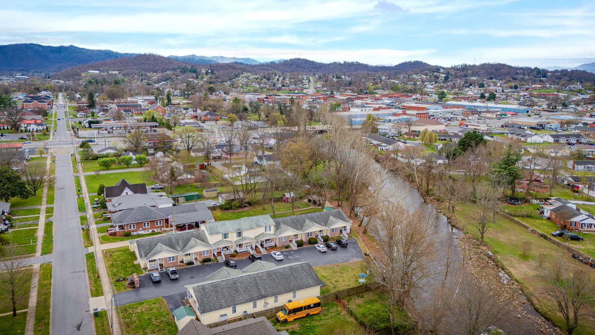 An aerial view of a town with houses, a school bus in a parking lot, a stream, and distant mountains under a cloudy sky.