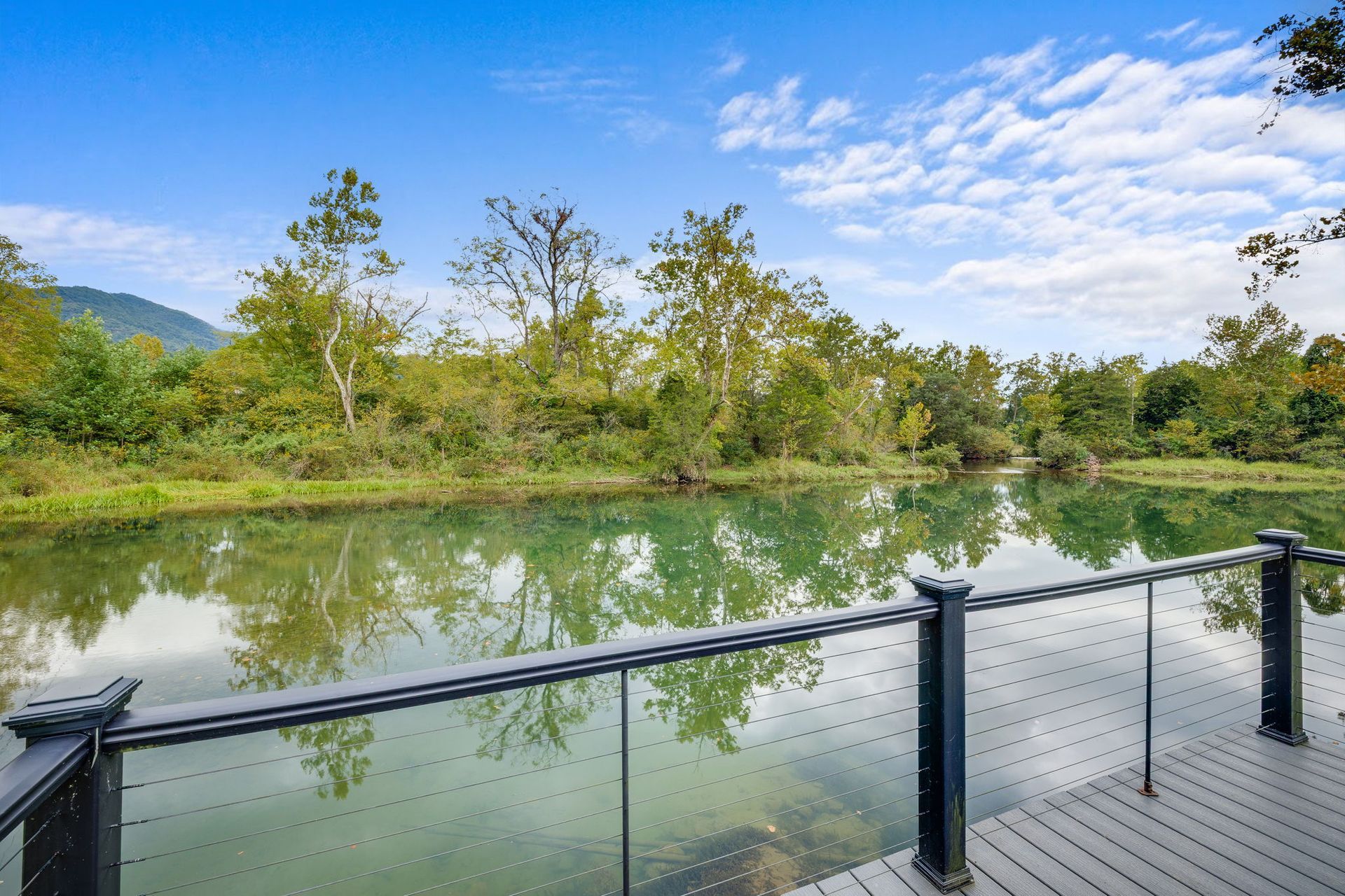 View of a pond from a deck with glass railing, surrounded by trees and a cloudy sky.
