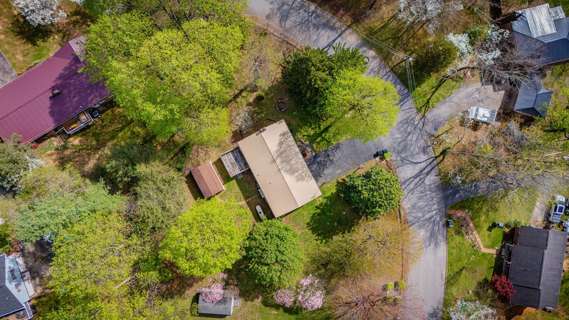 An aerial view of a wooded residential lot with a rectangular house, a shed, and a driveway winding through the trees.
