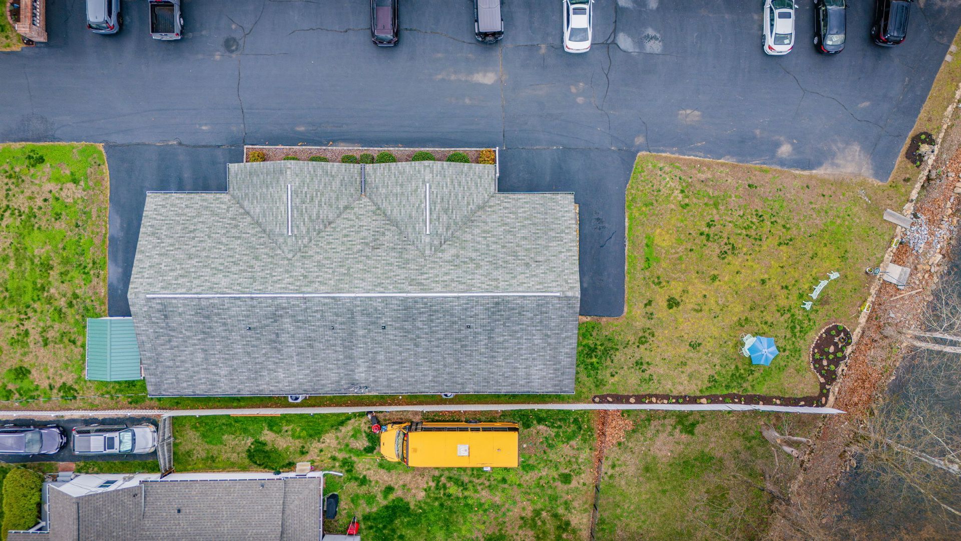 Aerial view of a parking lot, a gray-roofed building, a grassy lawn, and a yellow bus parked by a fence.