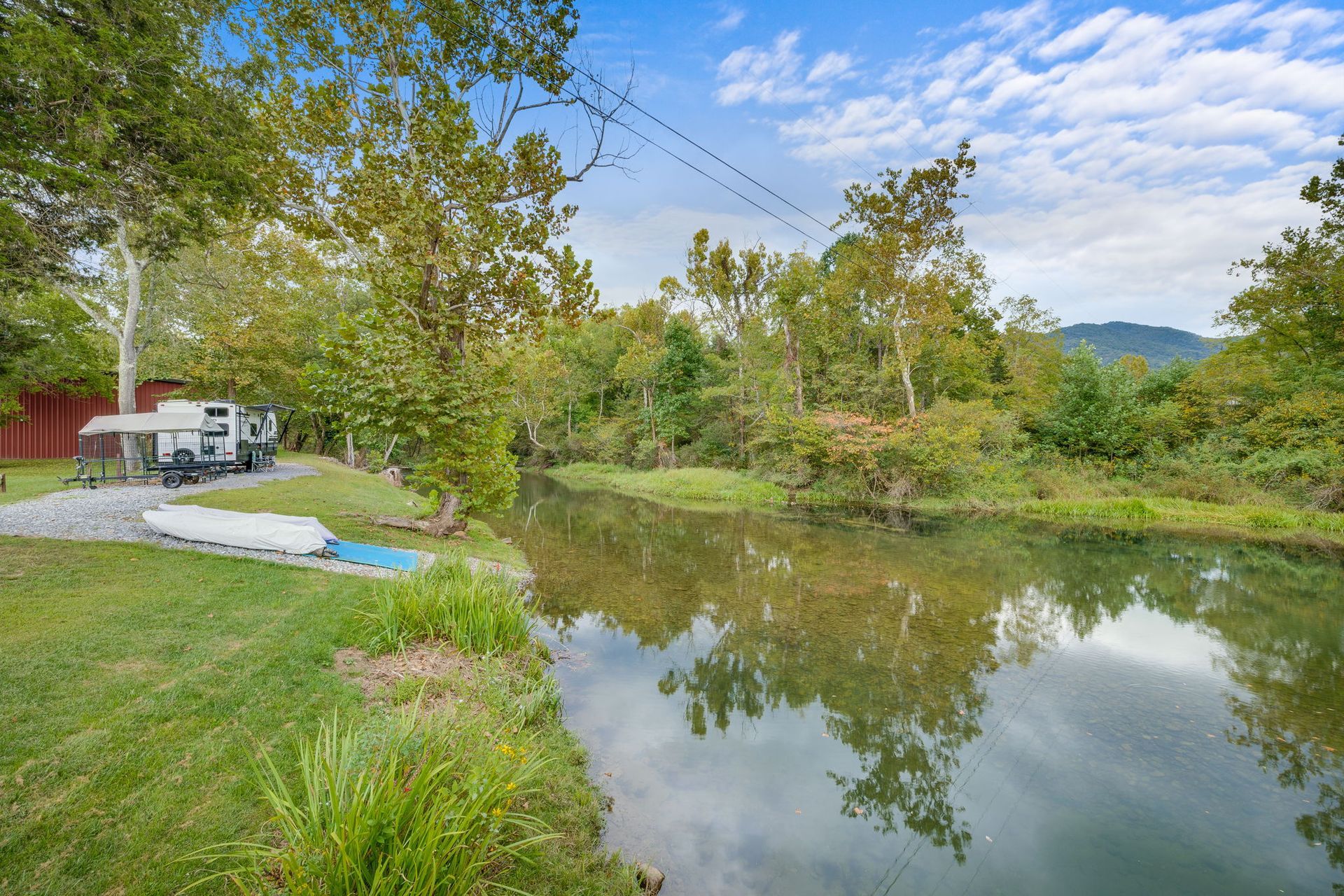 RV parked near a river on a grassy bank, surrounded by trees under a blue sky.
