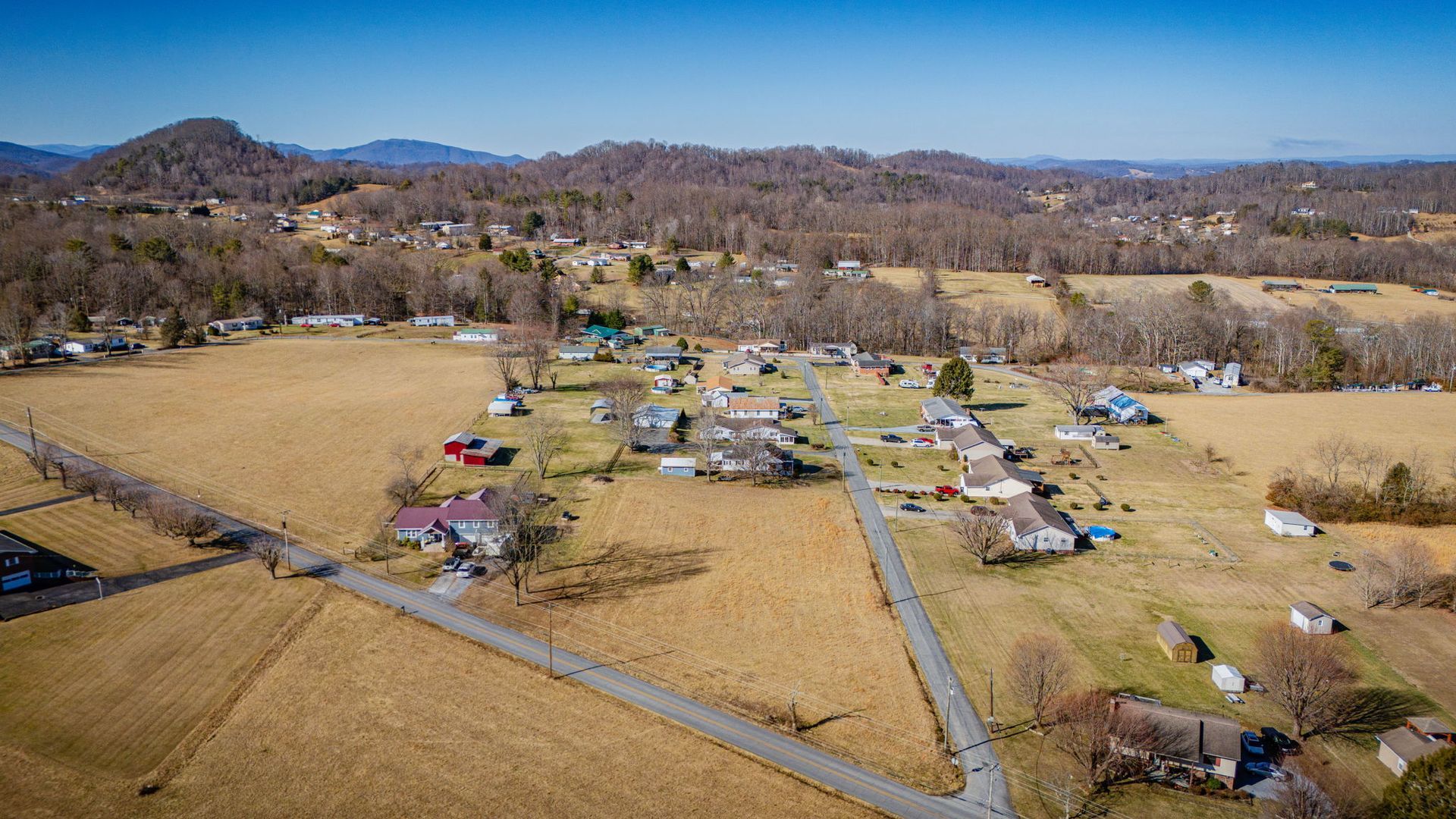 Aerial view of a rural residential area with houses, roads, fields, and forested hills under a blue sky.