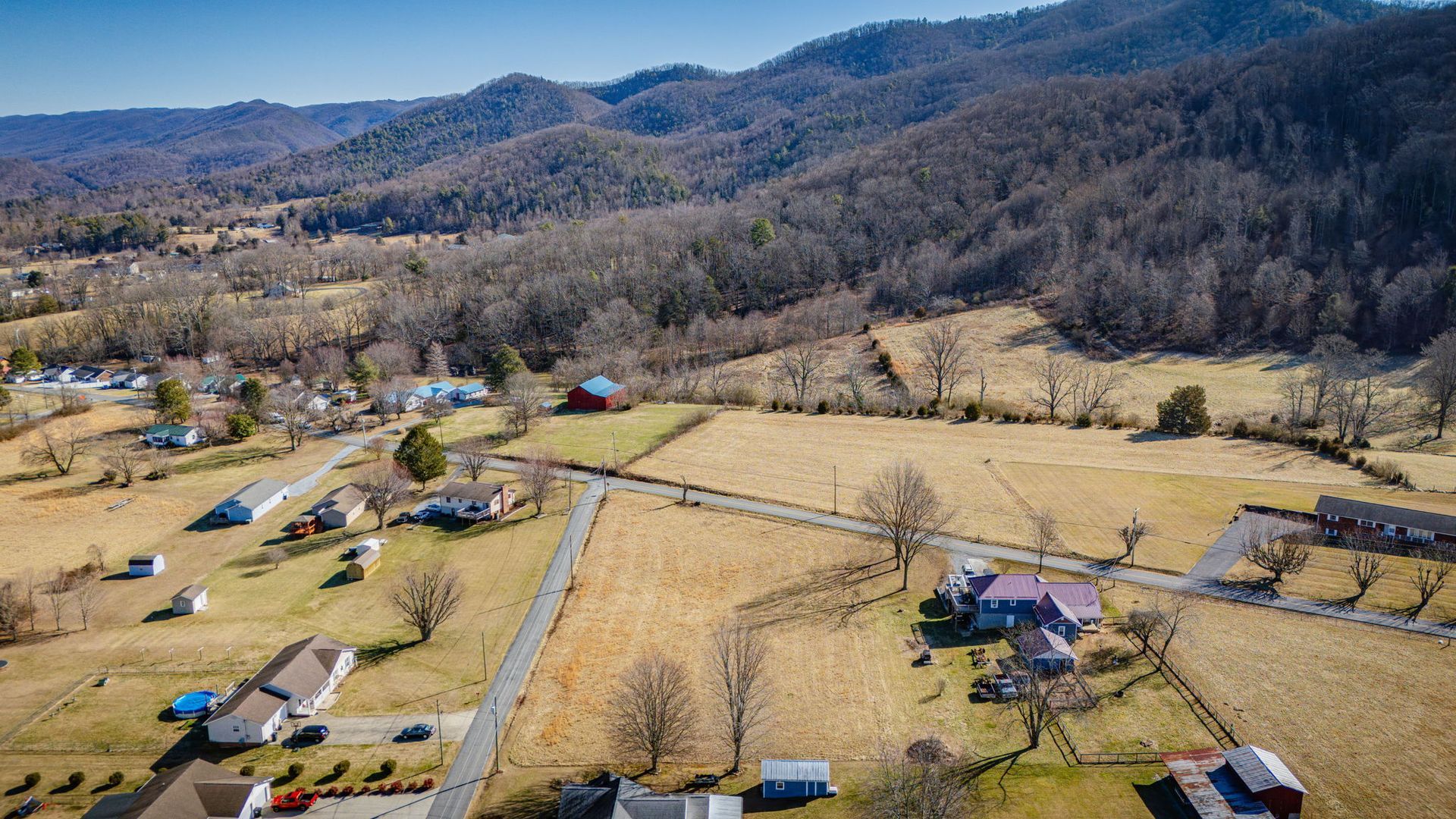 Aerial view of a rural landscape with houses, fields, roads, and wooded mountains under a blue sky.