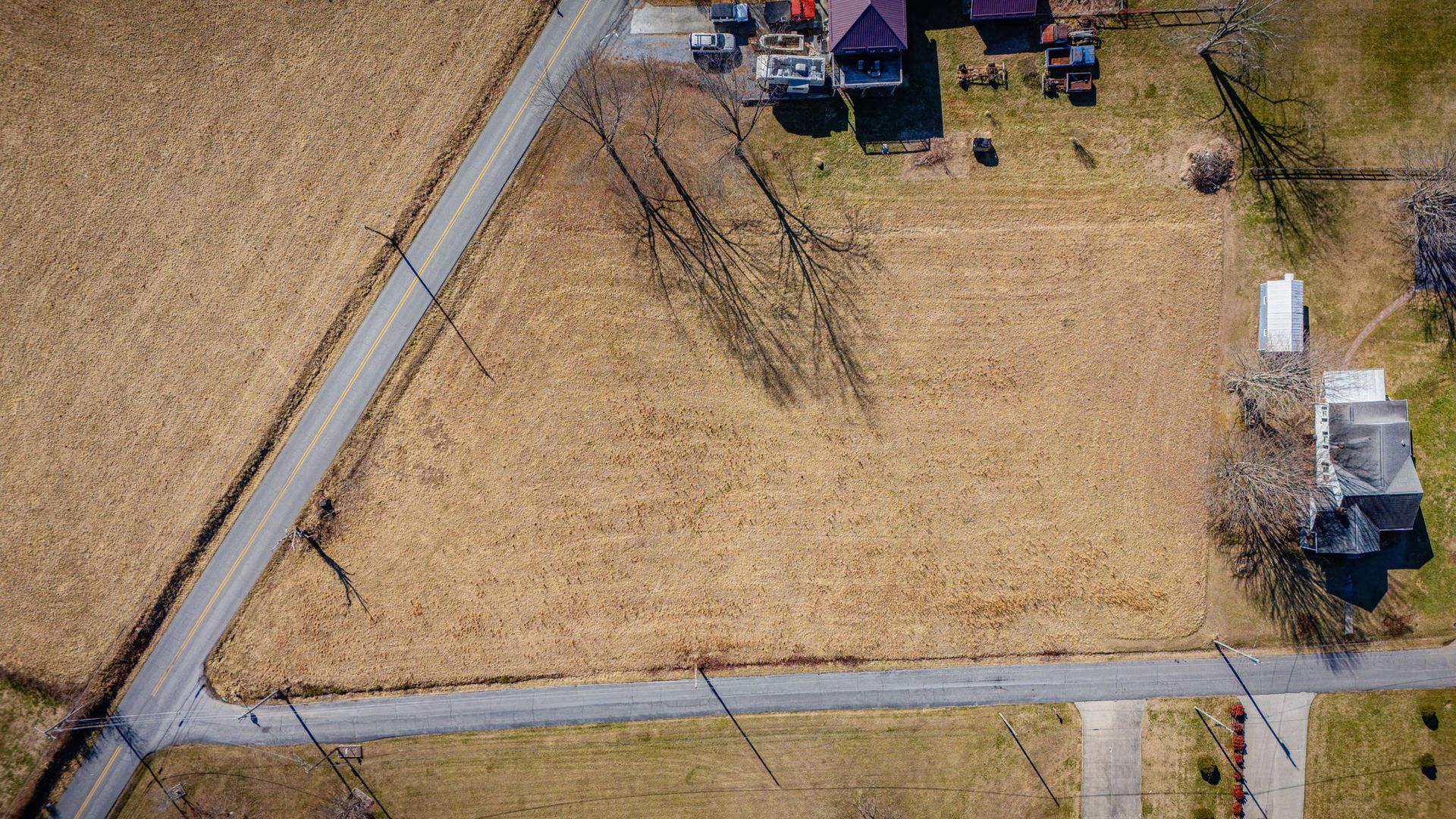 Aerial view of a vacant lot next to a road, surrounded by other houses and brown fields.