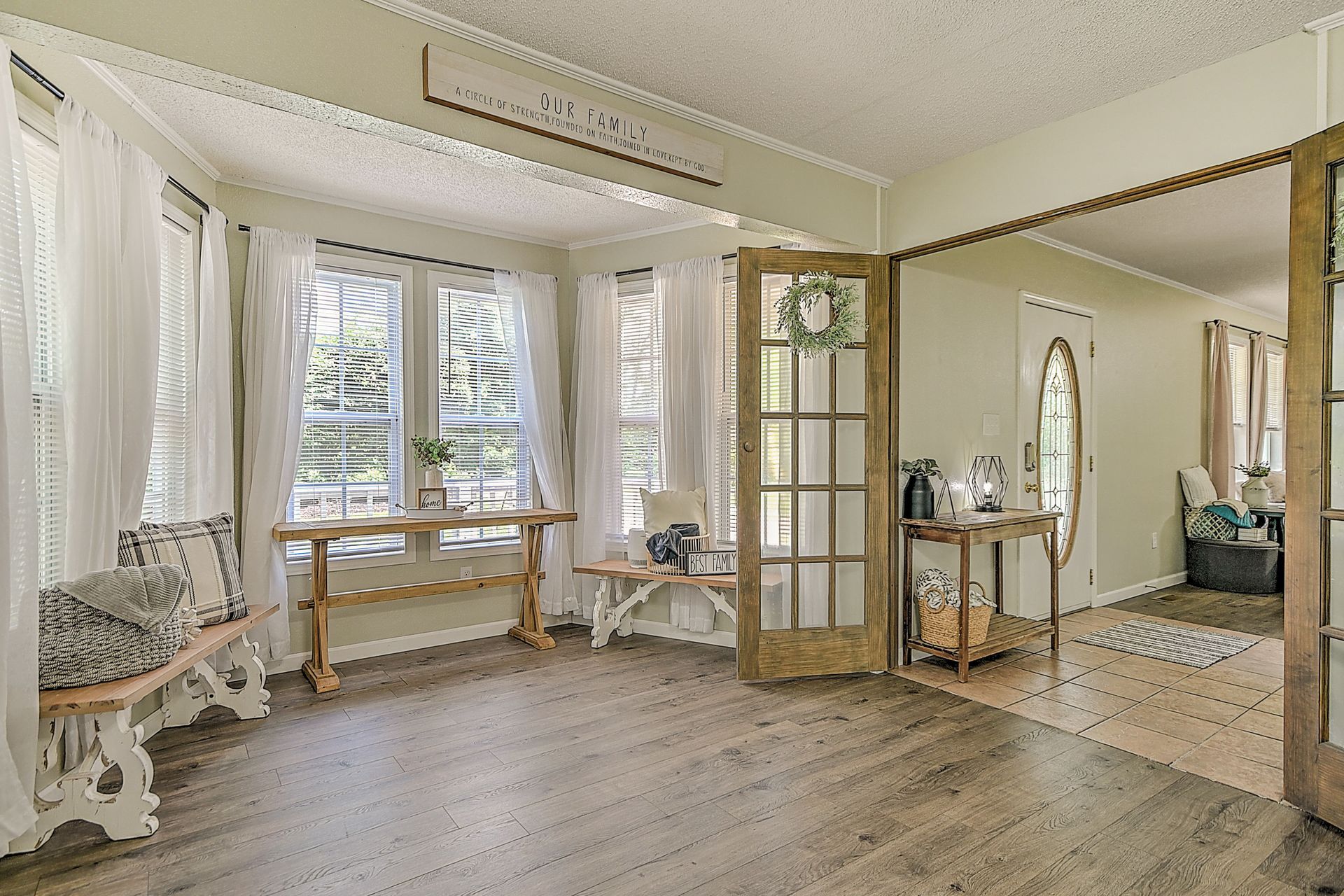 A living room with hardwood floors , white curtains , a table and a bench.