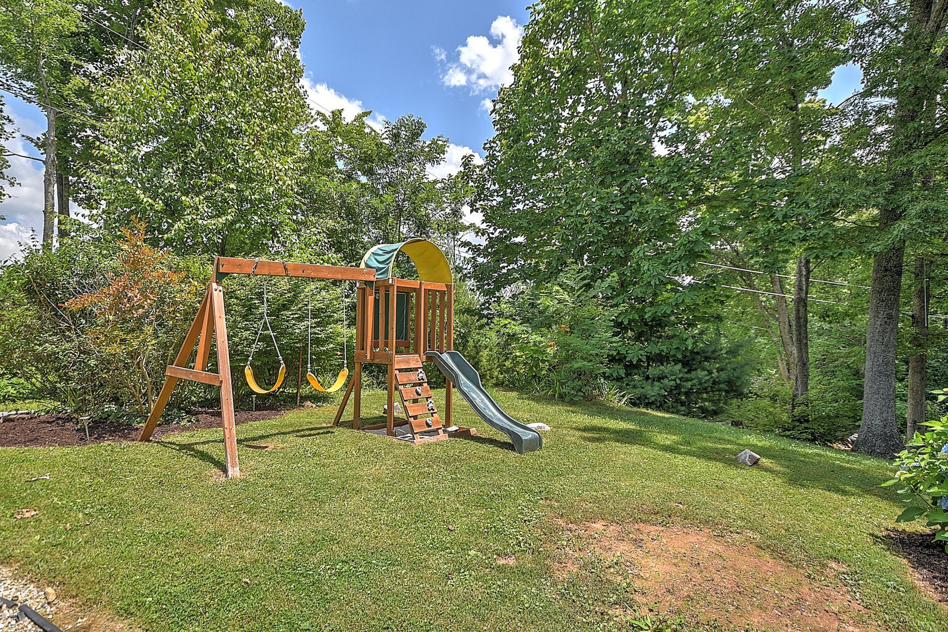A wooden playground with a slide and swings in a backyard surrounded by trees.