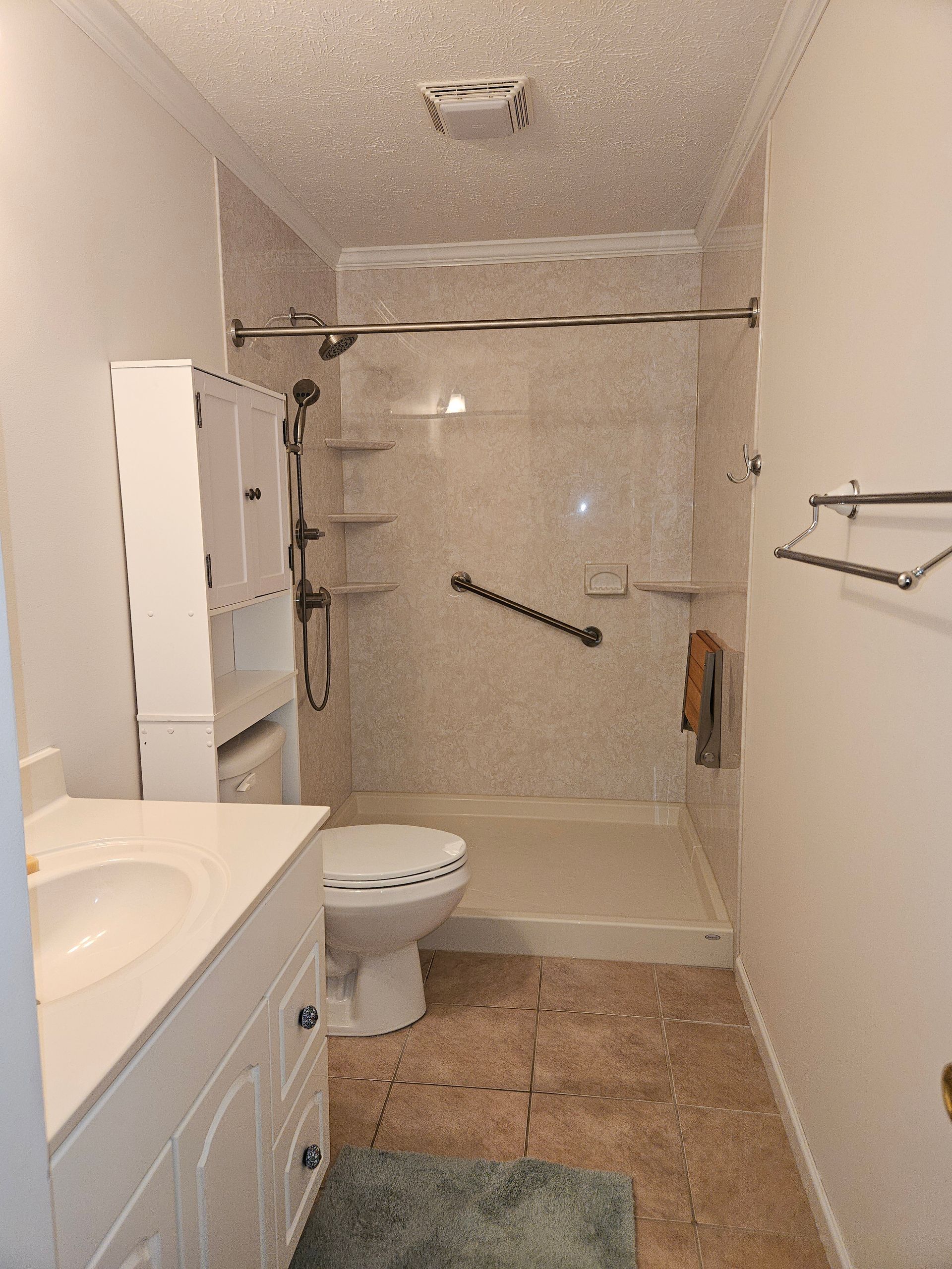 A narrow bathroom with a white vanity, a toilet, and a walk-in shower with grab bars and a beige tiled surround.