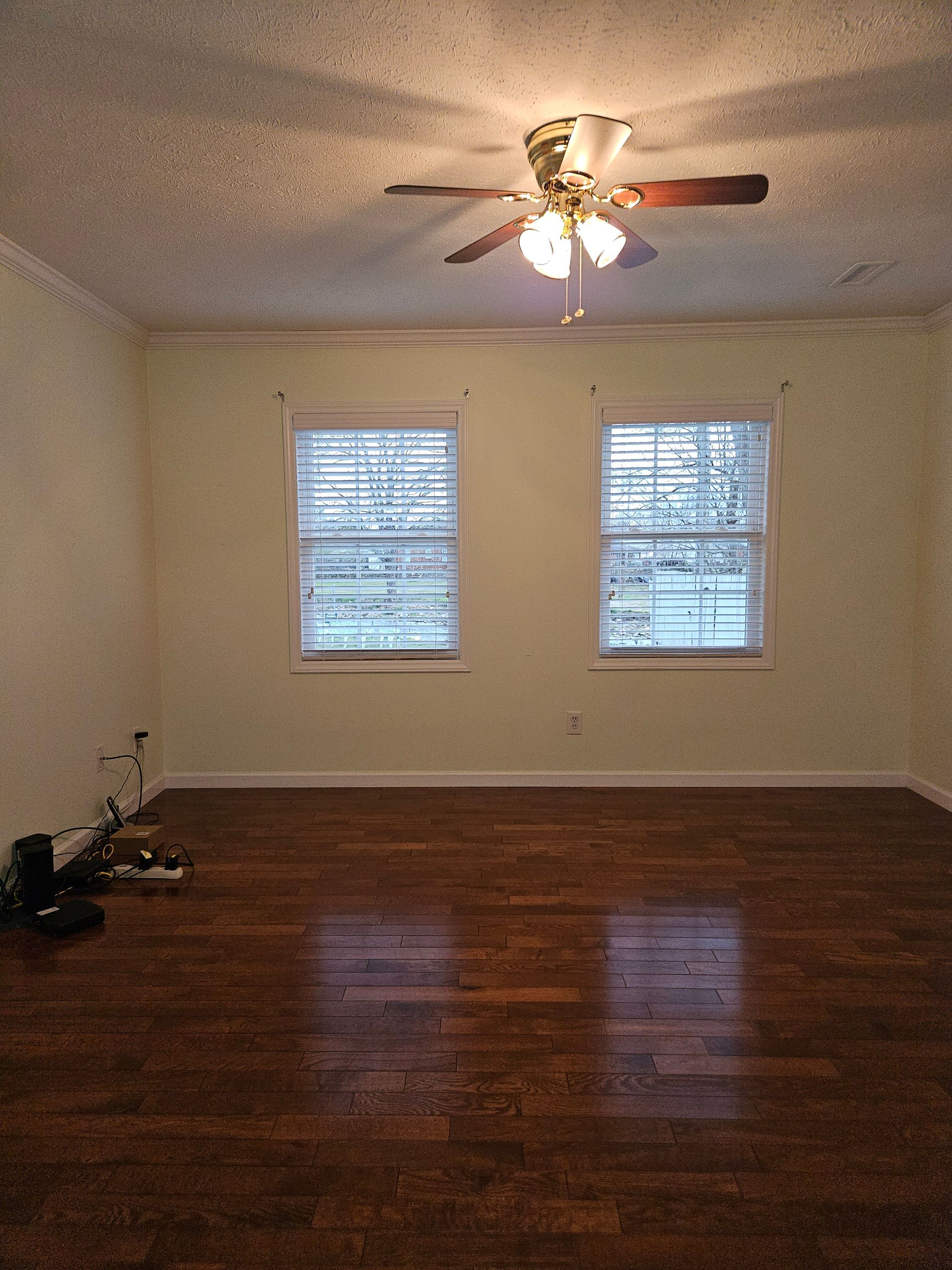 An empty room with light yellow walls, dark wood floors, two windows, and a ceiling fan.