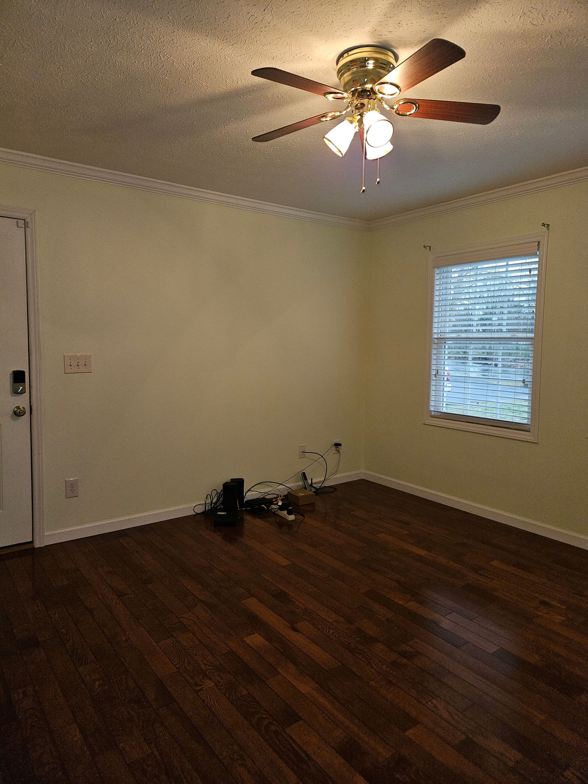 A bright, empty room with light walls, dark wood floors, a ceiling fan, and a window with blinds.