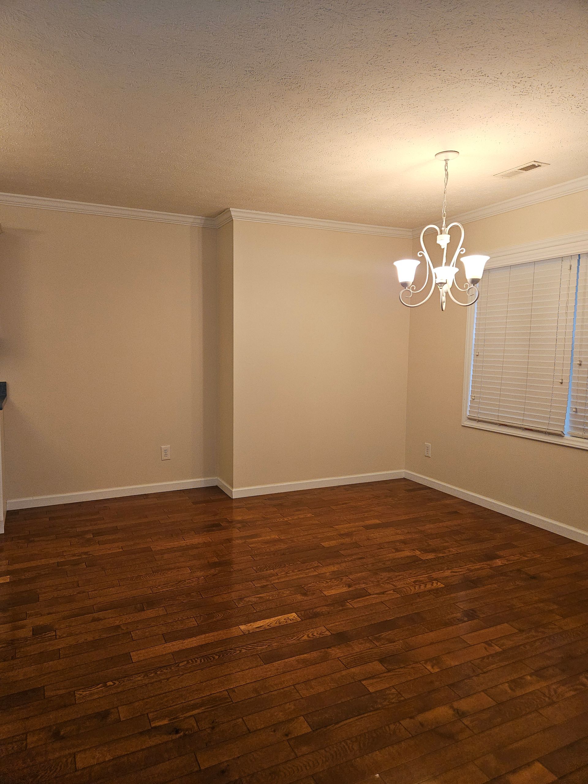 An empty dining room with dark wood floors, beige walls, white crown molding, and a traditional white chandelier.