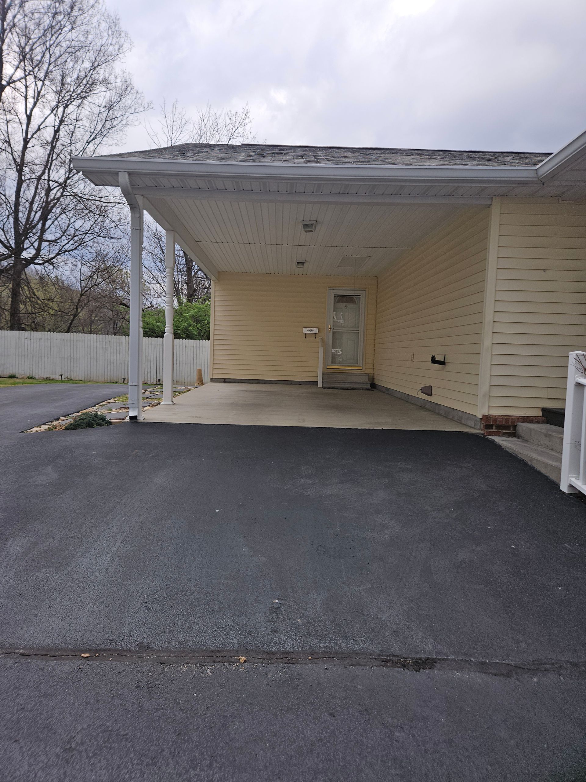 A light yellow house with a concrete driveway leading to a covered carport area under a cloudy sky.