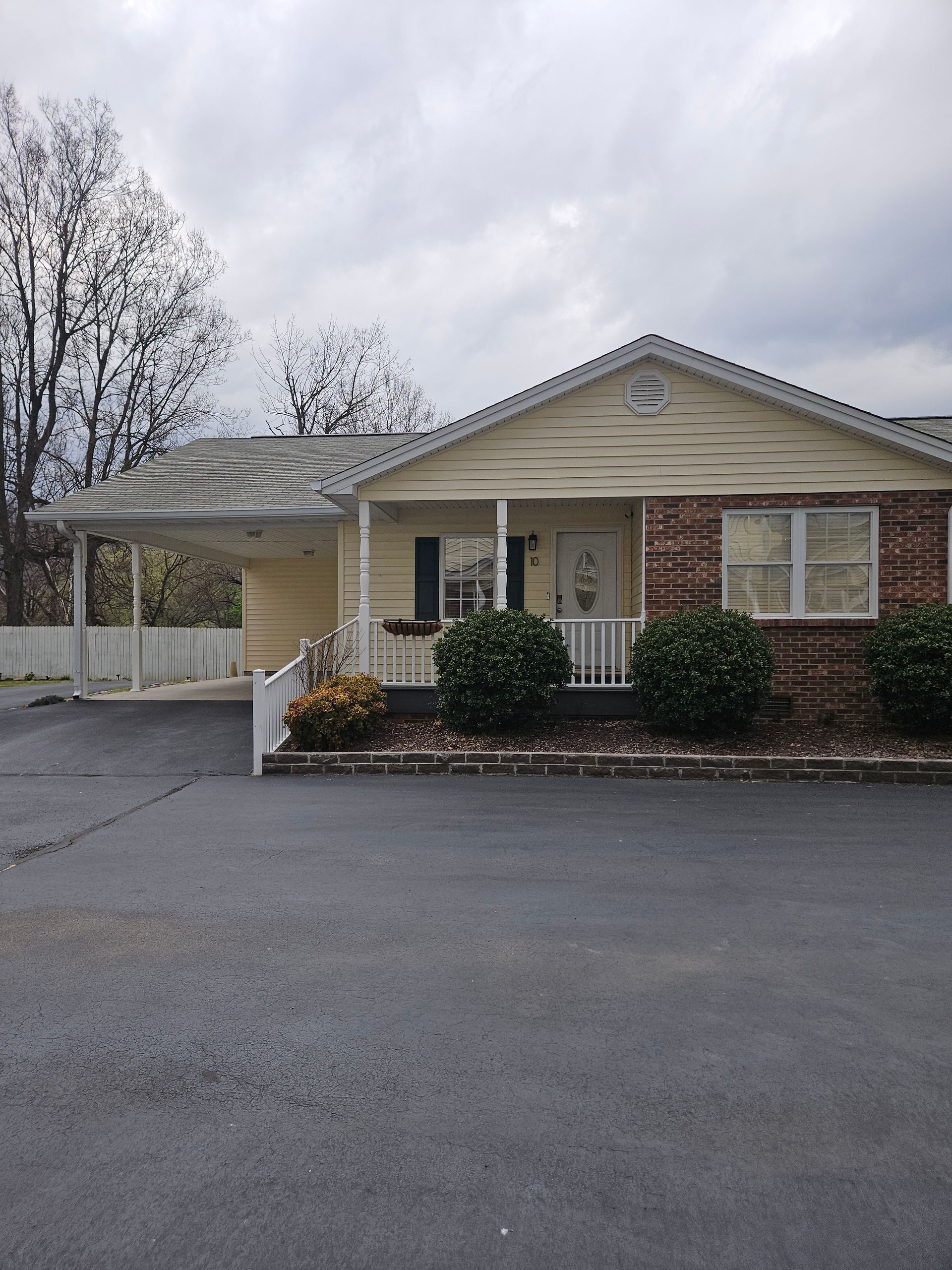 A single-story, light yellow house with a brick exterior, a front porch, and a side carport under a cloudy sky.