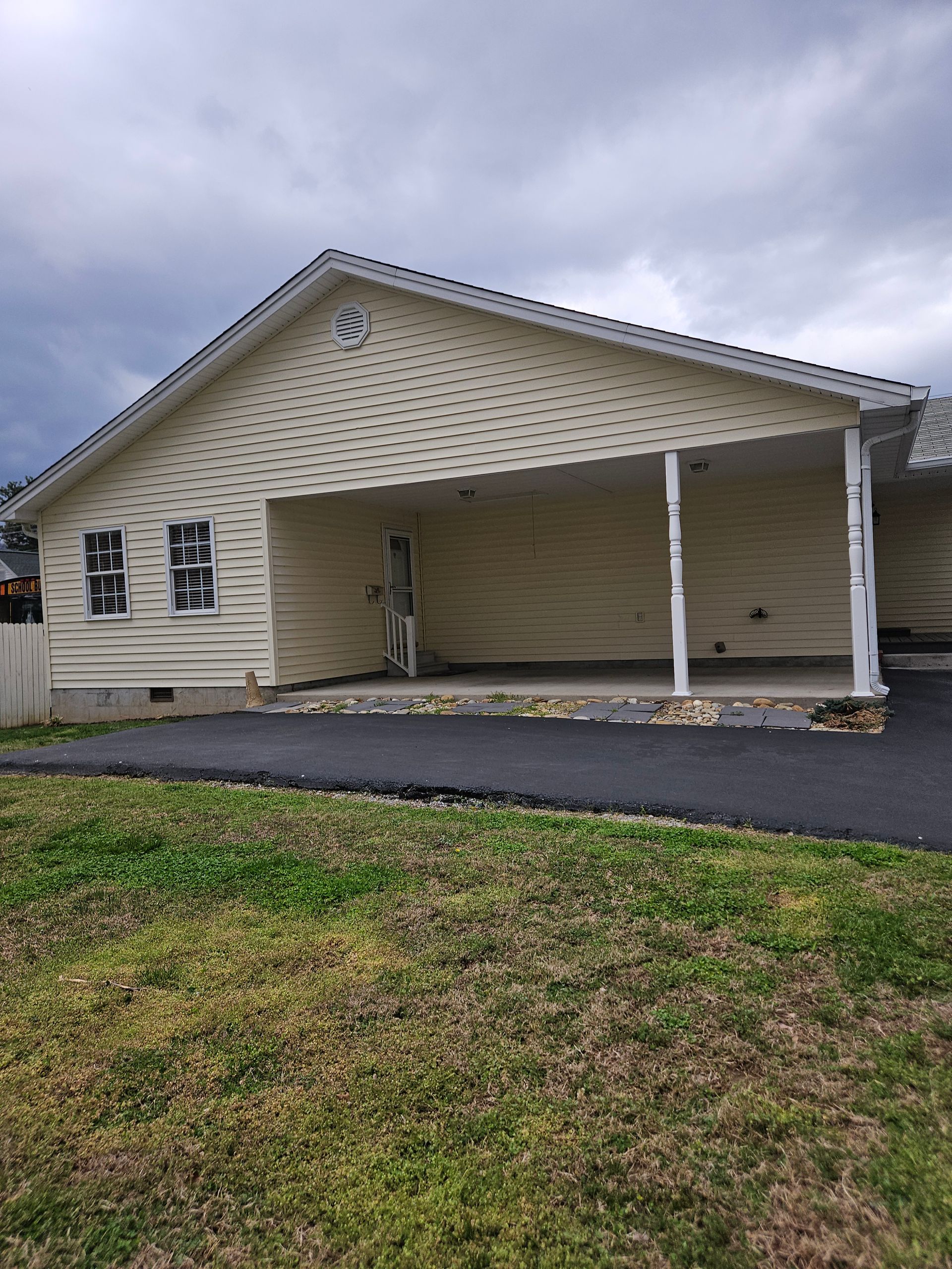 Light-yellow house with a paved driveway and a covered front porch featuring two white columns under a cloudy sky.