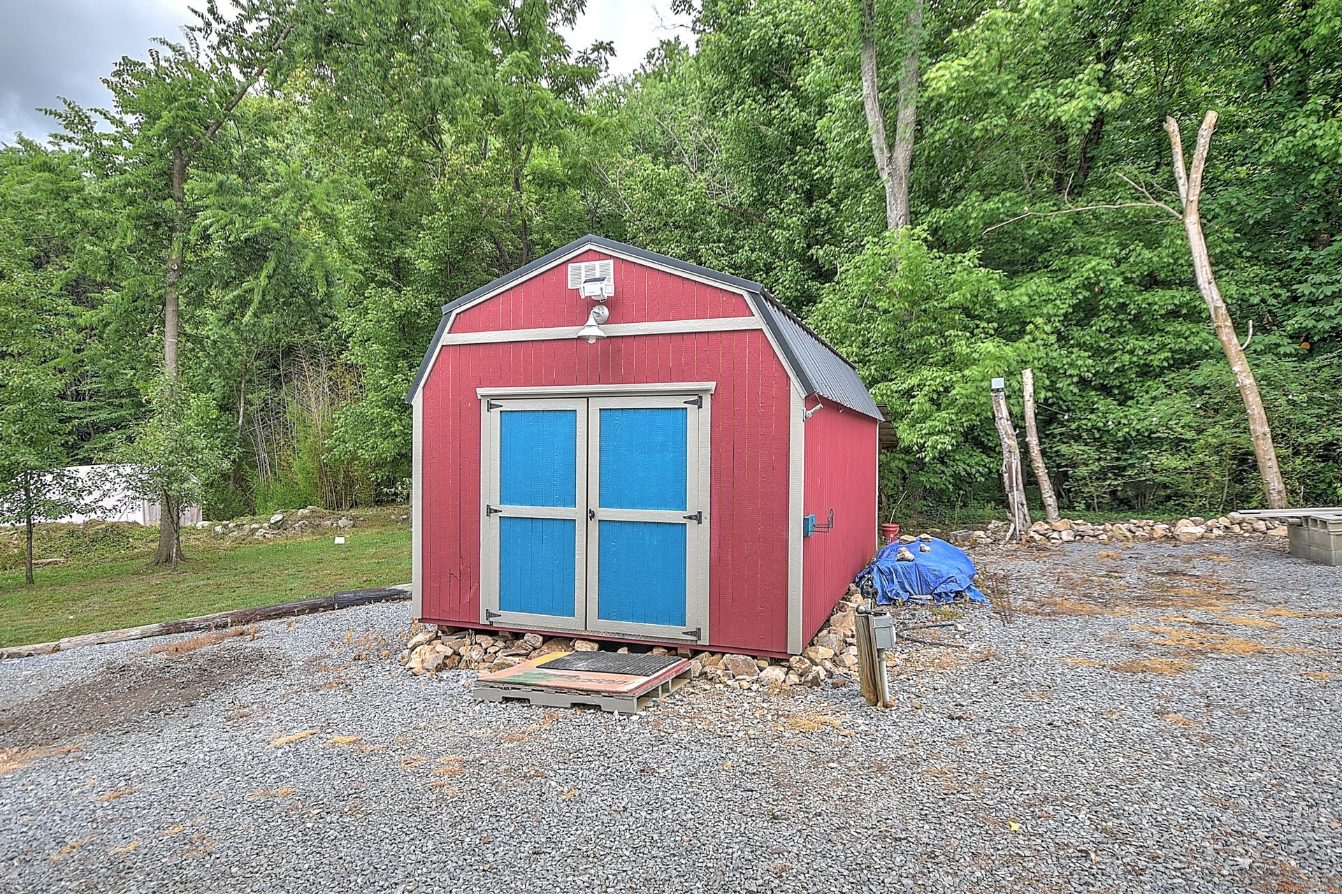 Red shed with blue double doors on a gravel lot, surrounded by green trees.