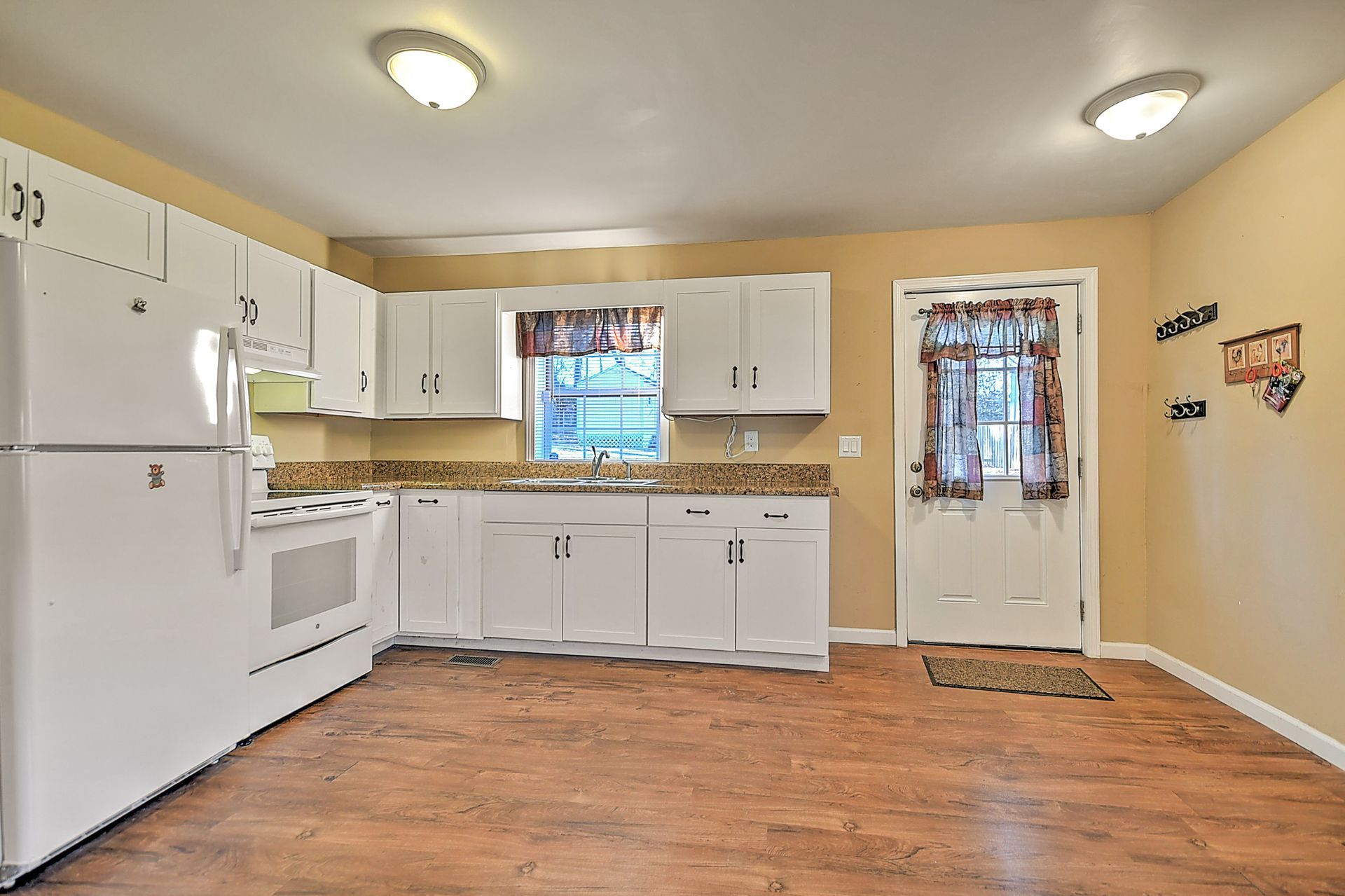 Bright kitchen with white cabinets, appliances, and tan walls. Wooden floor. Door to the outside.