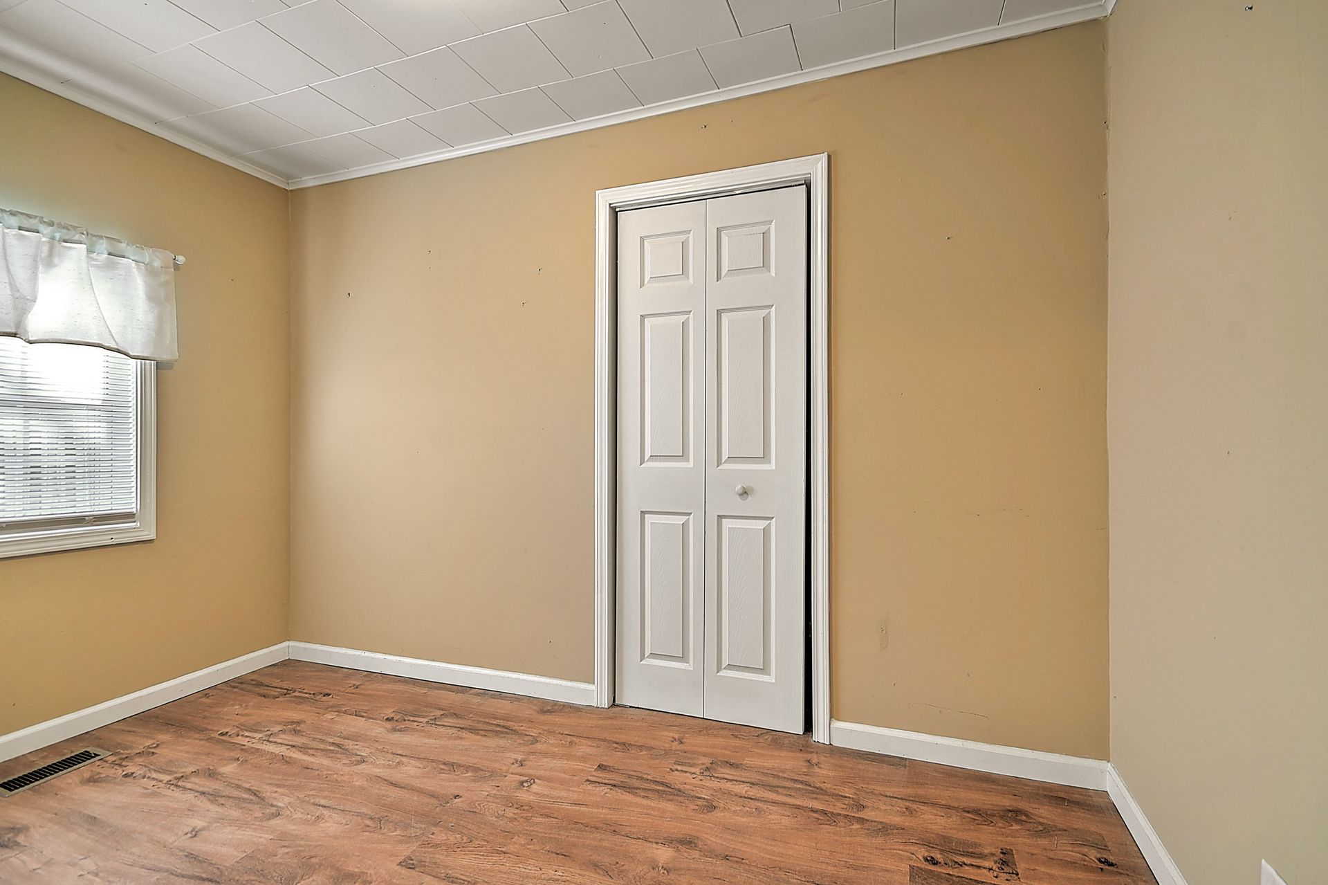 Empty room with tan walls, a white closet door, window with a curtain, and wood-look floor.