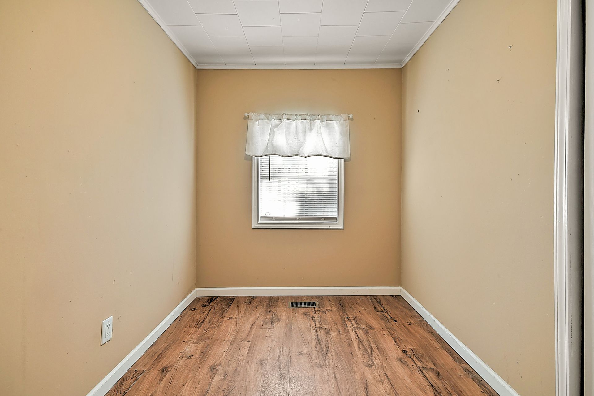 Empty room with wood-look floor, tan walls, small window with white curtain, and white trim.
