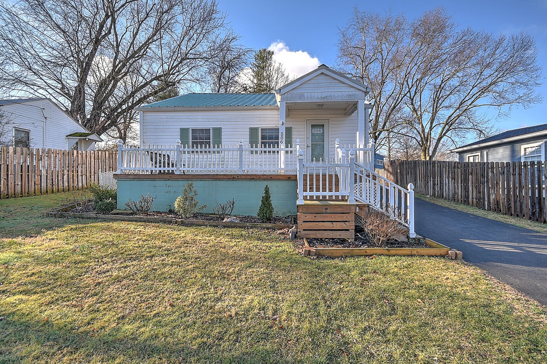 Small white house with porch, green metal roof, fenced yard, and driveway.