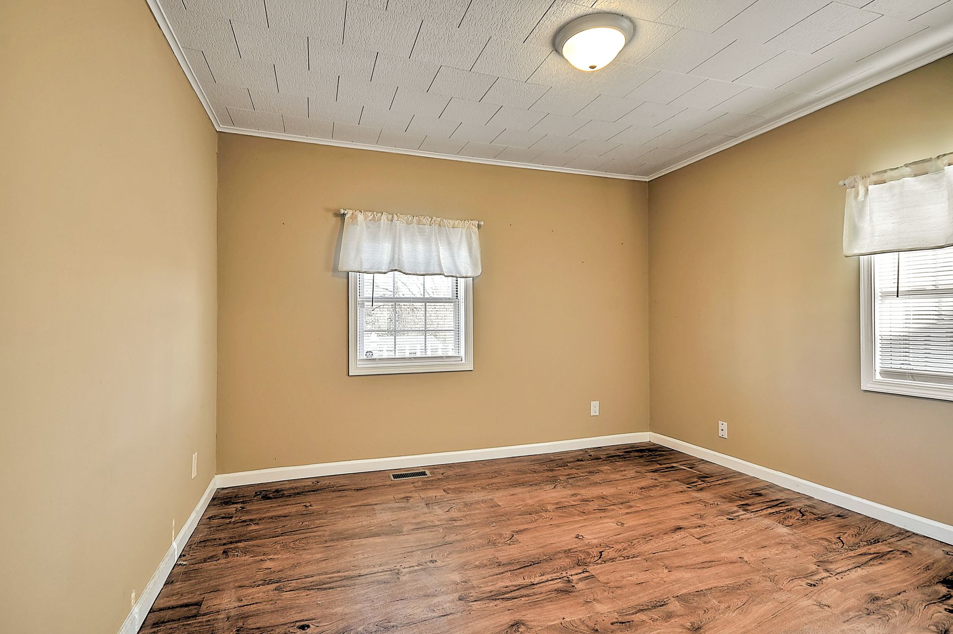 Empty room with tan walls, white ceiling and trim, two windows with curtains, and wooden floor.