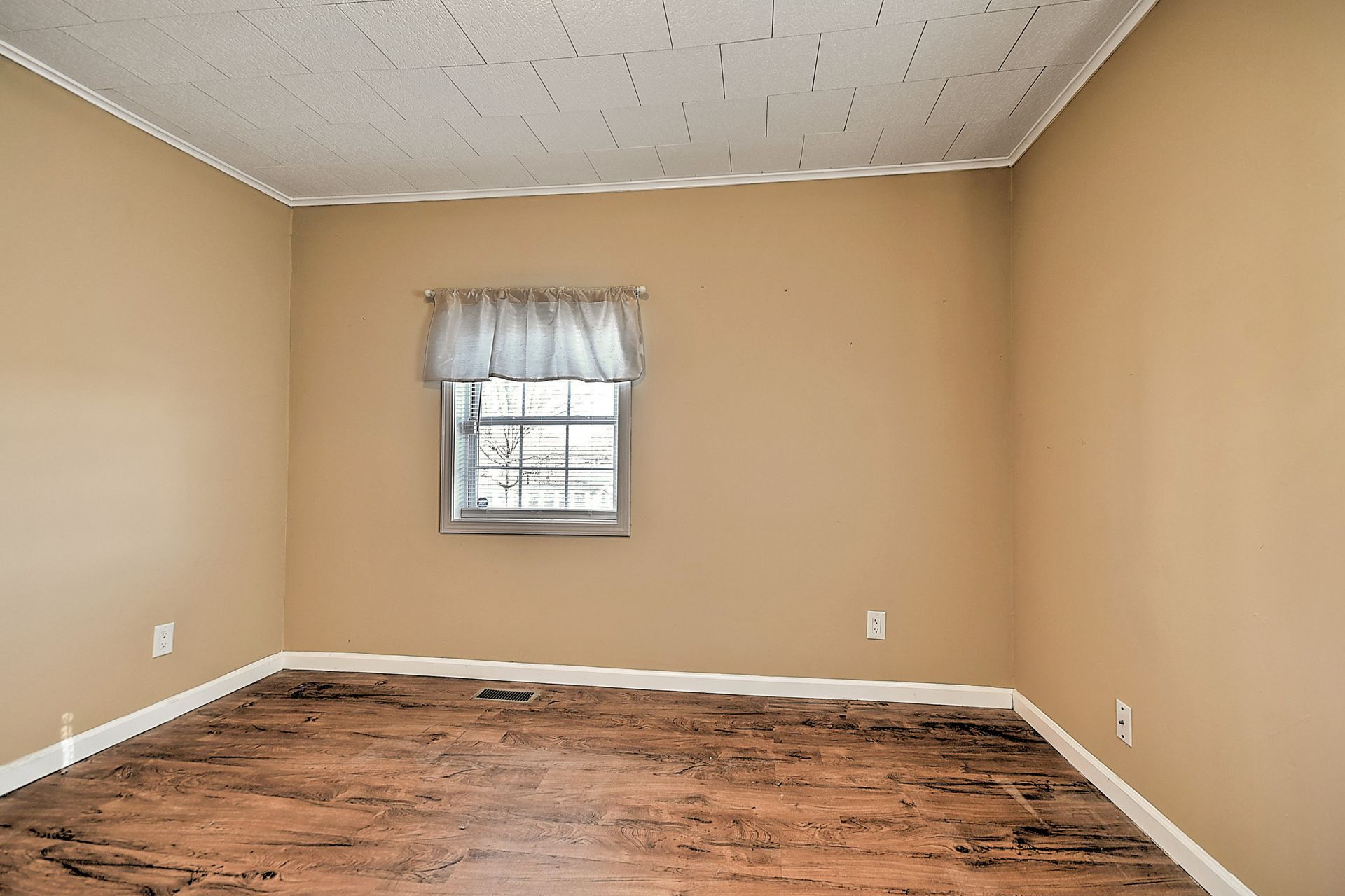 Empty room with beige walls, a small window with a curtain, and damaged brown flooring.
